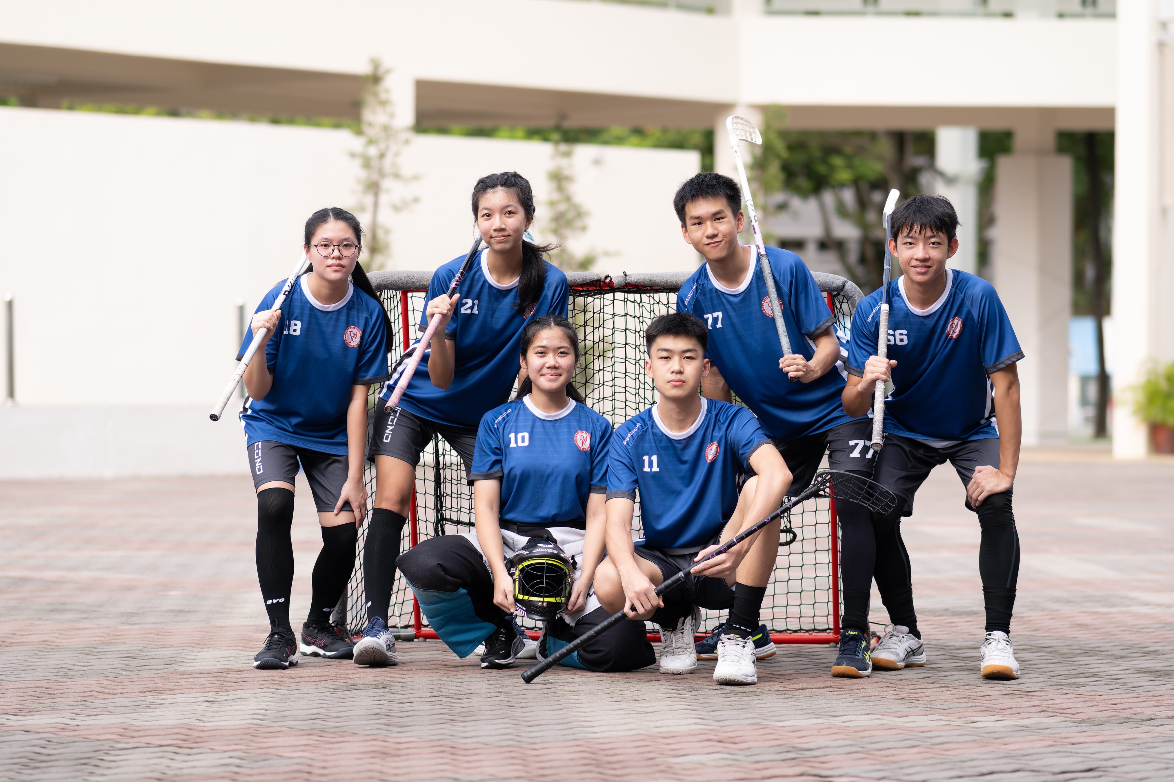 Six junior college students in blue numbered floorball jerseys pose with their sticks in front of a floorball goal outdoors, with two seated in front holding a goalkeeper's helmet.