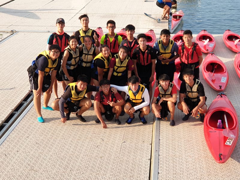 A group of young people wearing life jackets pose on a dock beside several red kayaks near water.