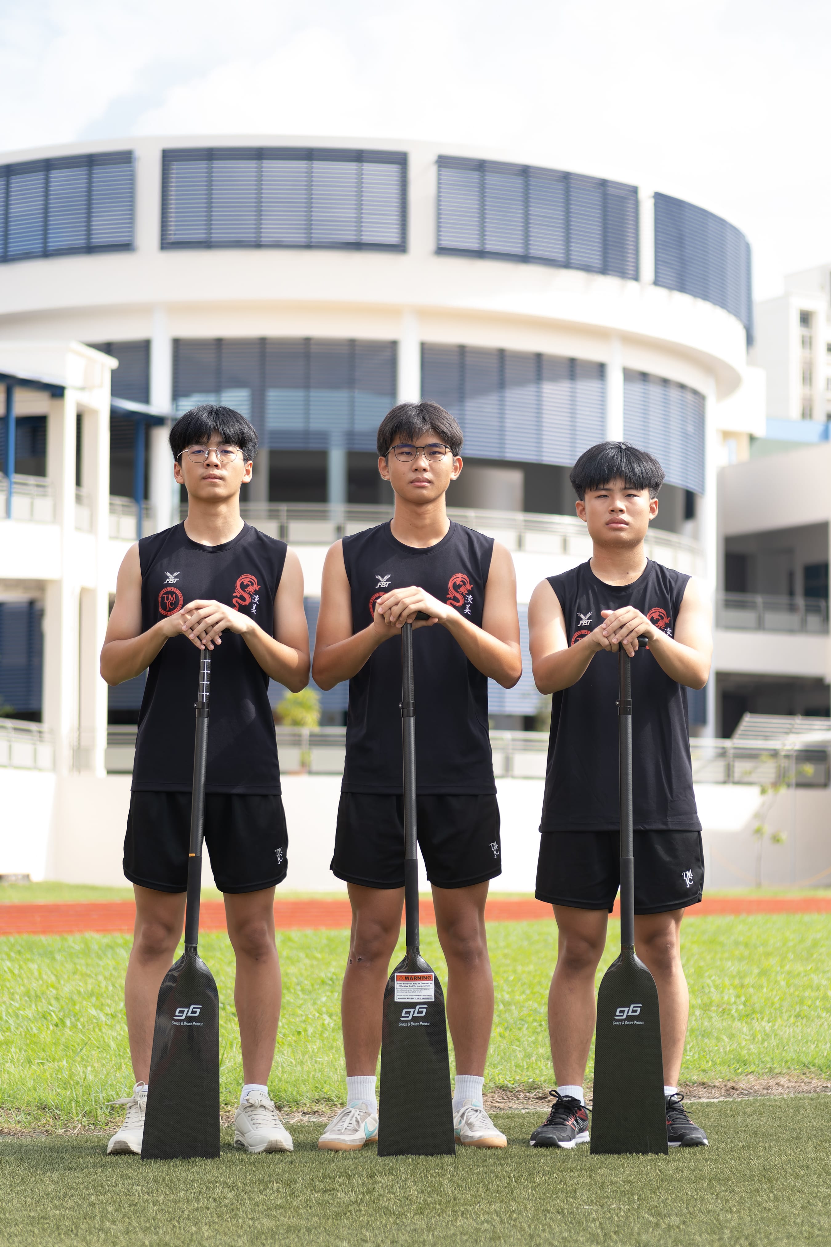 Three TMJC male students in black sleeveless dragon boat jerseys stand side by side on a grass field, each resting both hands on an upright dragon boat paddle. A curved school building is visible behind them.