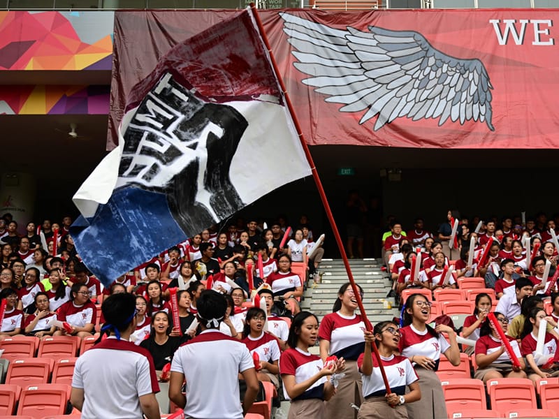 Students in red and white uniforms cheer in stadium stands as one waves a large dark flag.
