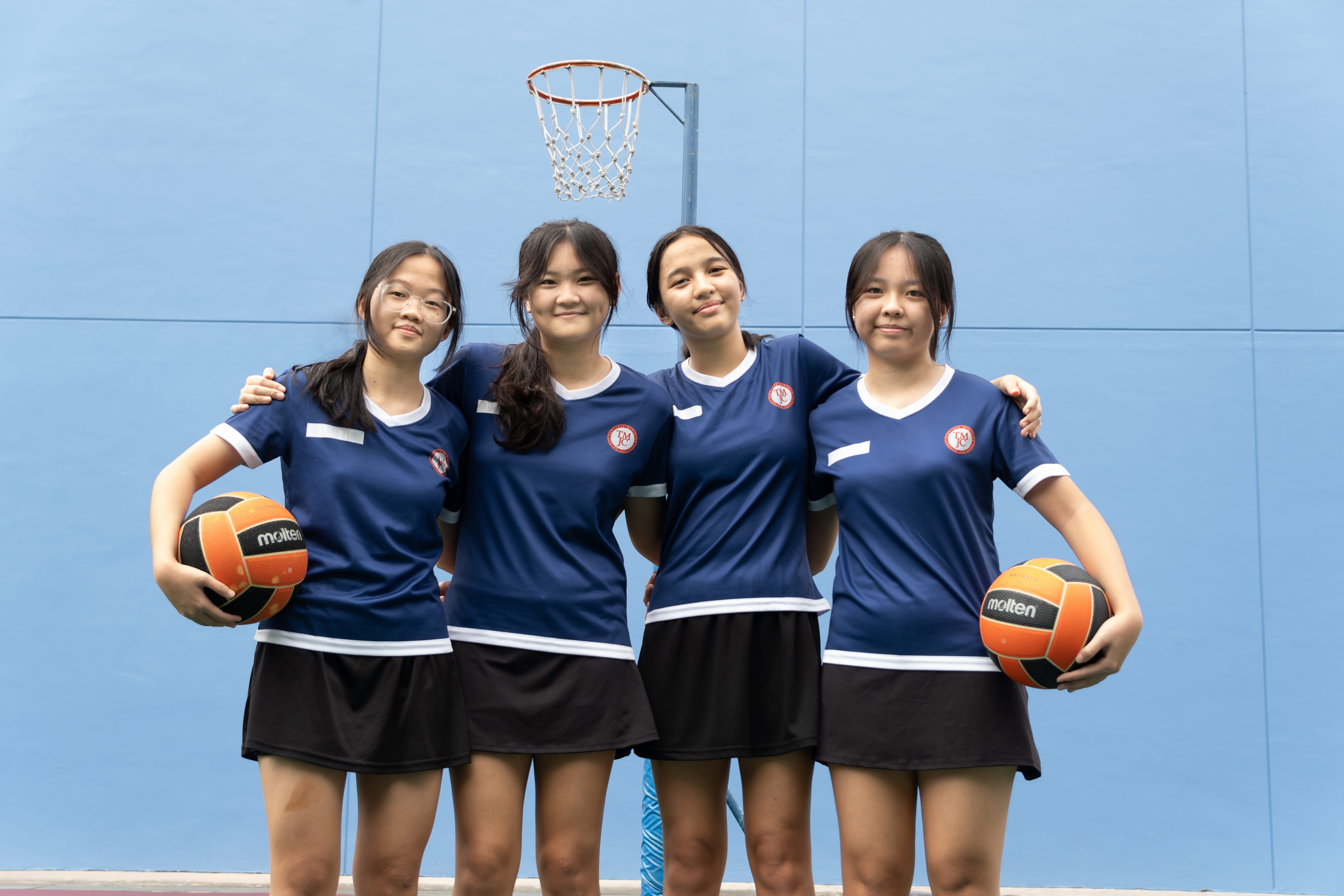 Four TMJC female netball players in navy blue jerseys and black skirts stand arm-in-arm in front of a netball hoop, smiling at the camera. Two of them hold orange and black Molten netballs.