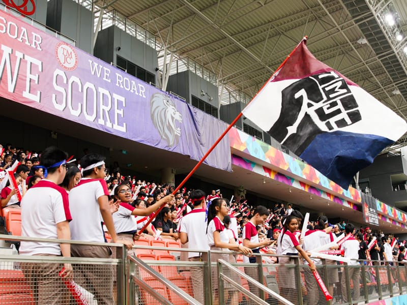 Students in white and red uniforms cheer in stadium stands beneath a banner reading "We Roar We Score" as a flag waves.
