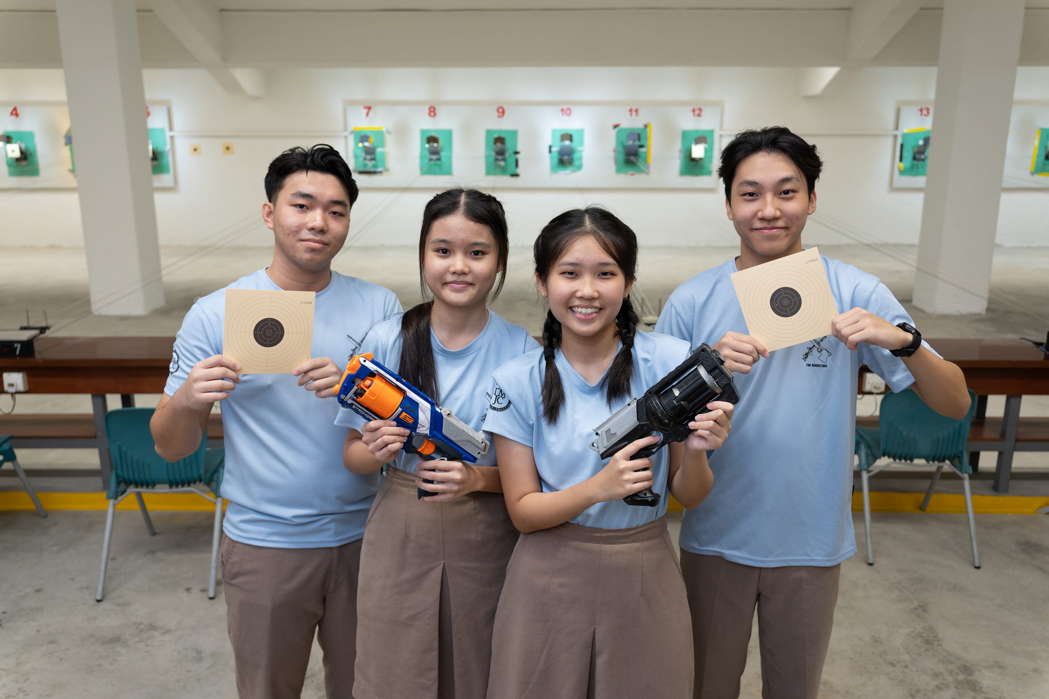 Four students in light blue TMJC uniforms smile at the camera inside a shooting range, holding target sheets and air pistols, with numbered target lanes visible in the background.