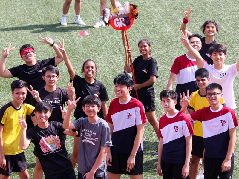 A group of smiling students on a green field, making peace signs, with one holding a bird mascot on a stick.