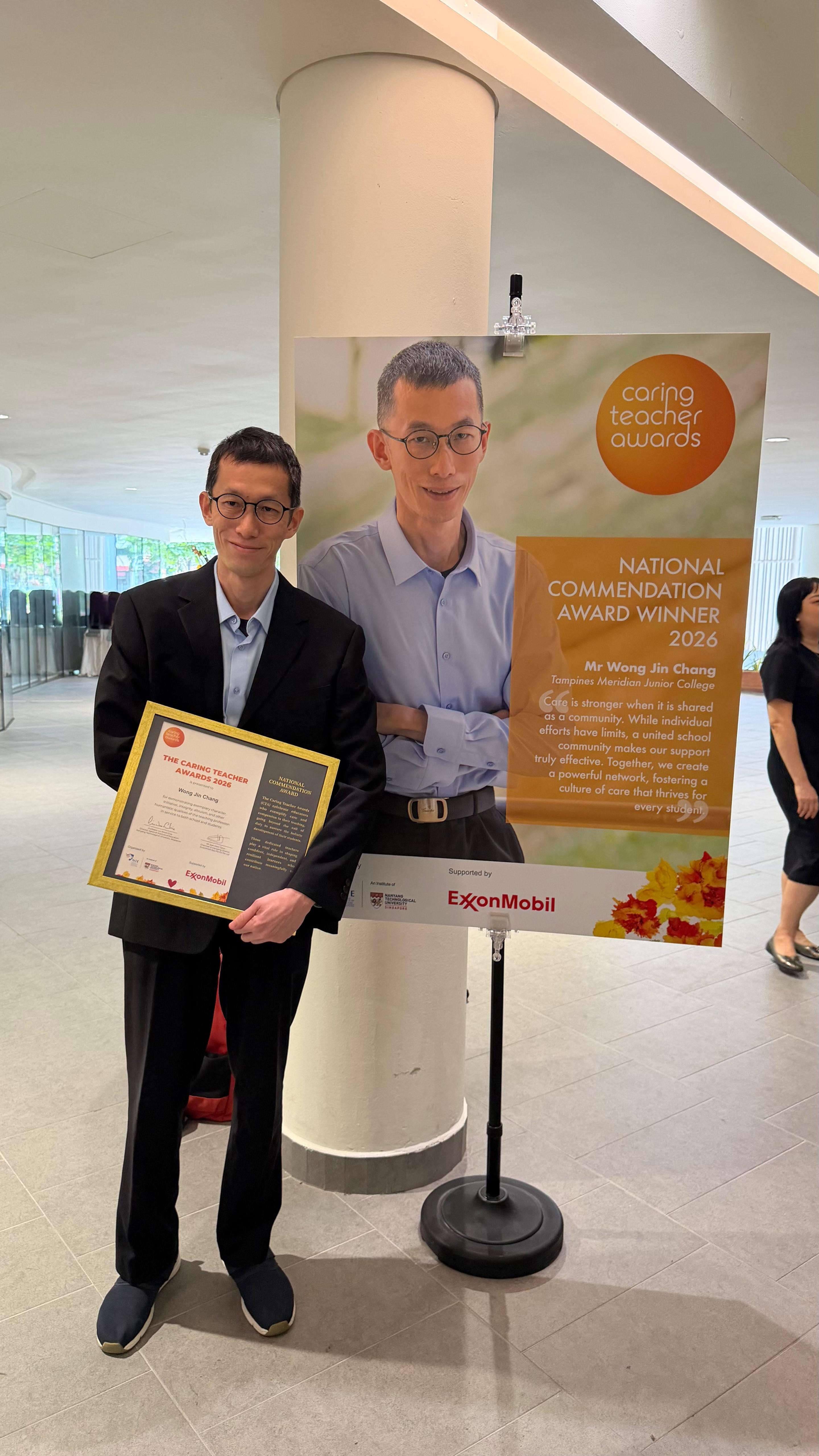 Mr Wong Jin Chang, a teacher from Tampines Meridian Junior College, stands holding his framed Caring Teacher Awards 2026 National Commendation Award certificate, next to a banner displaying his photo and a quote about fostering a culture of care in schools. The award is supported by ExxonMobil.