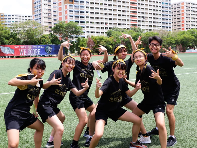 Eight students in matching black Olympus shirts and yellow headbands pose playfully on a green sports field.