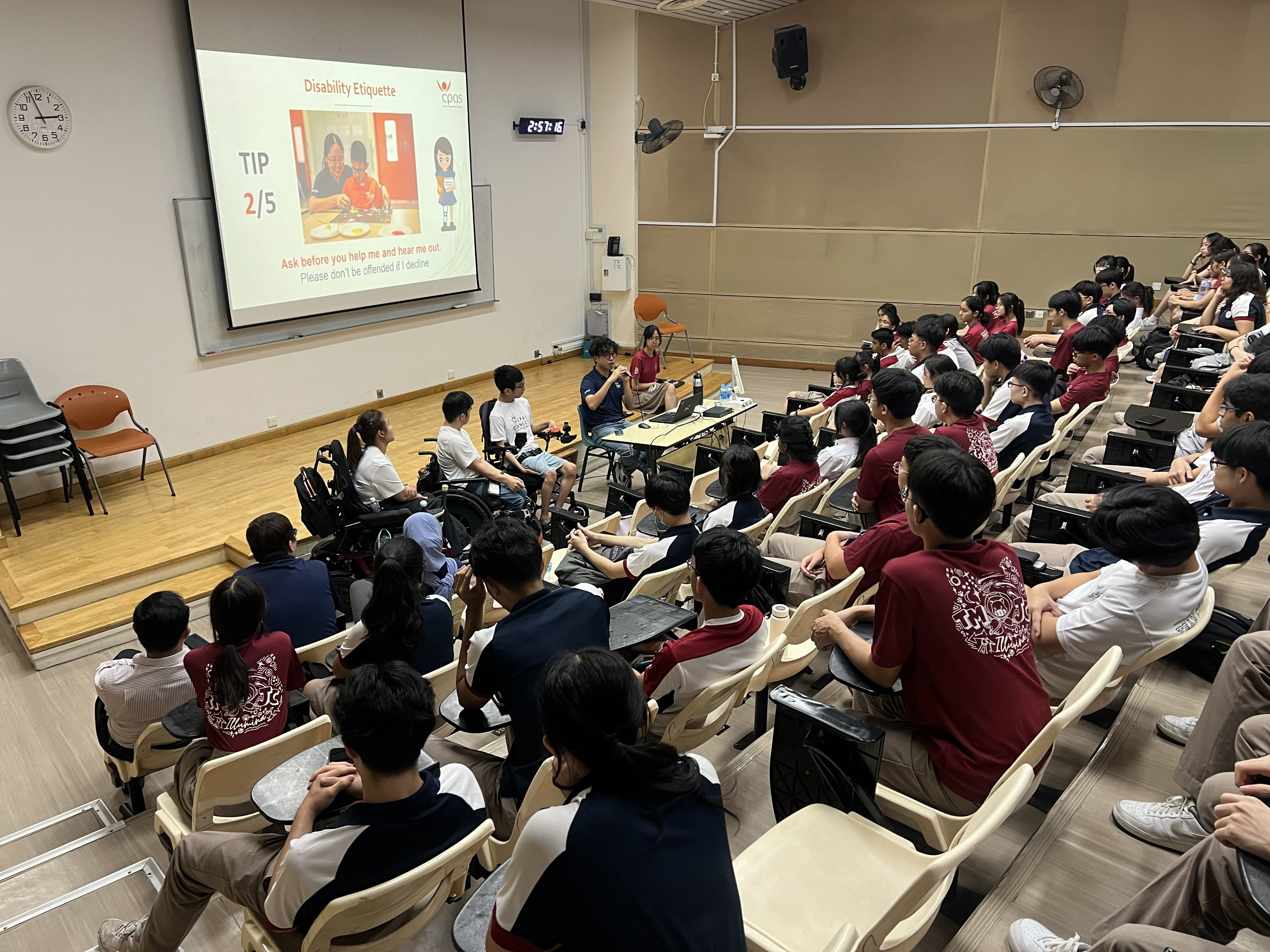 Students fill a lecture theatre watching a presentation on "Disability Etiquette" by CPAS. On stage, a speaker in a wheelchair and another student present to the audience. The slide on screen reads: "Ask before you help me and hear me out. Please don't be offended if I decline."