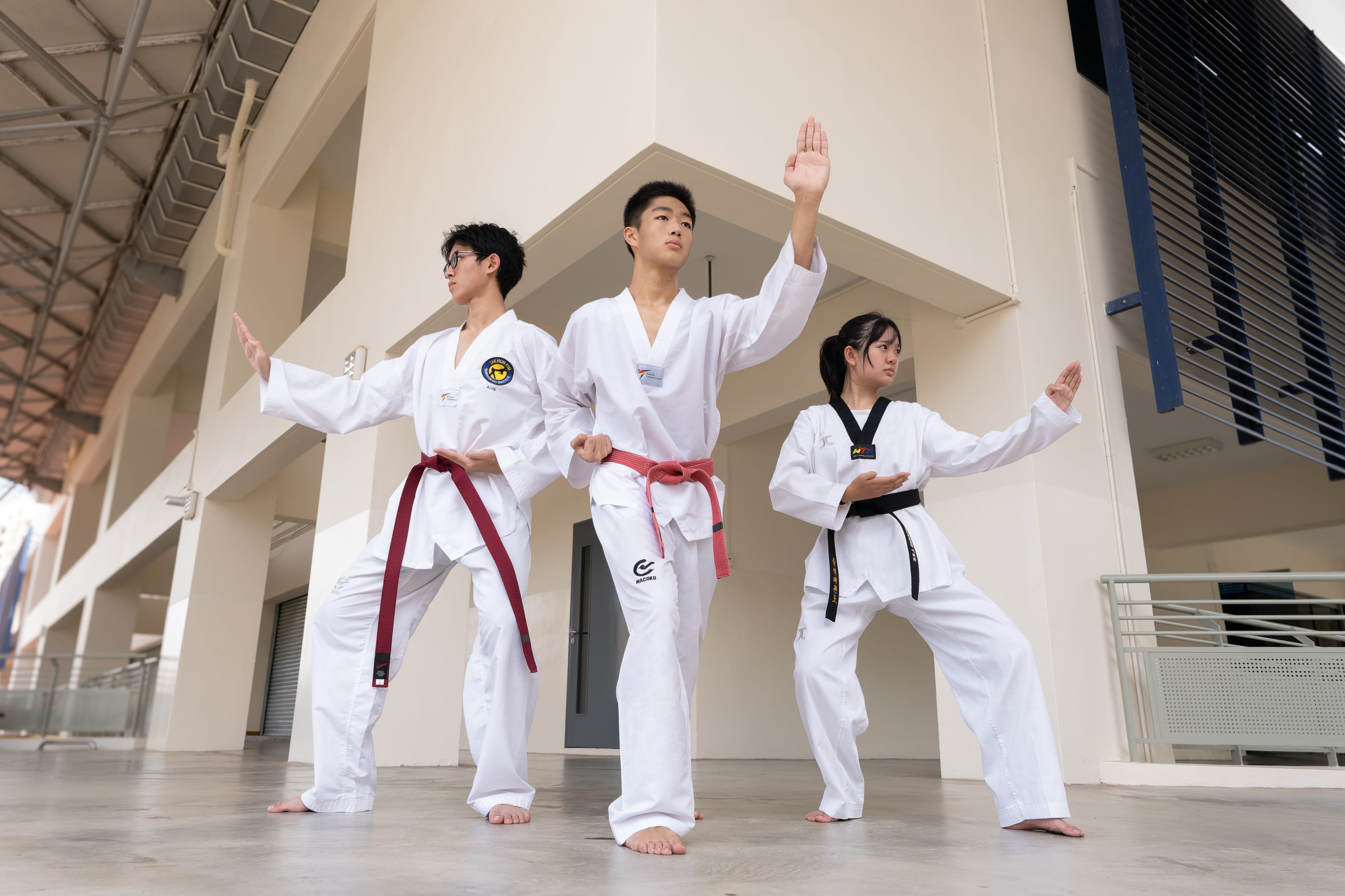 Three taekwondo students in white uniforms — two wearing red belts and one wearing a black belt — performing a synchronised poomsae stance outdoors at a covered walkway.