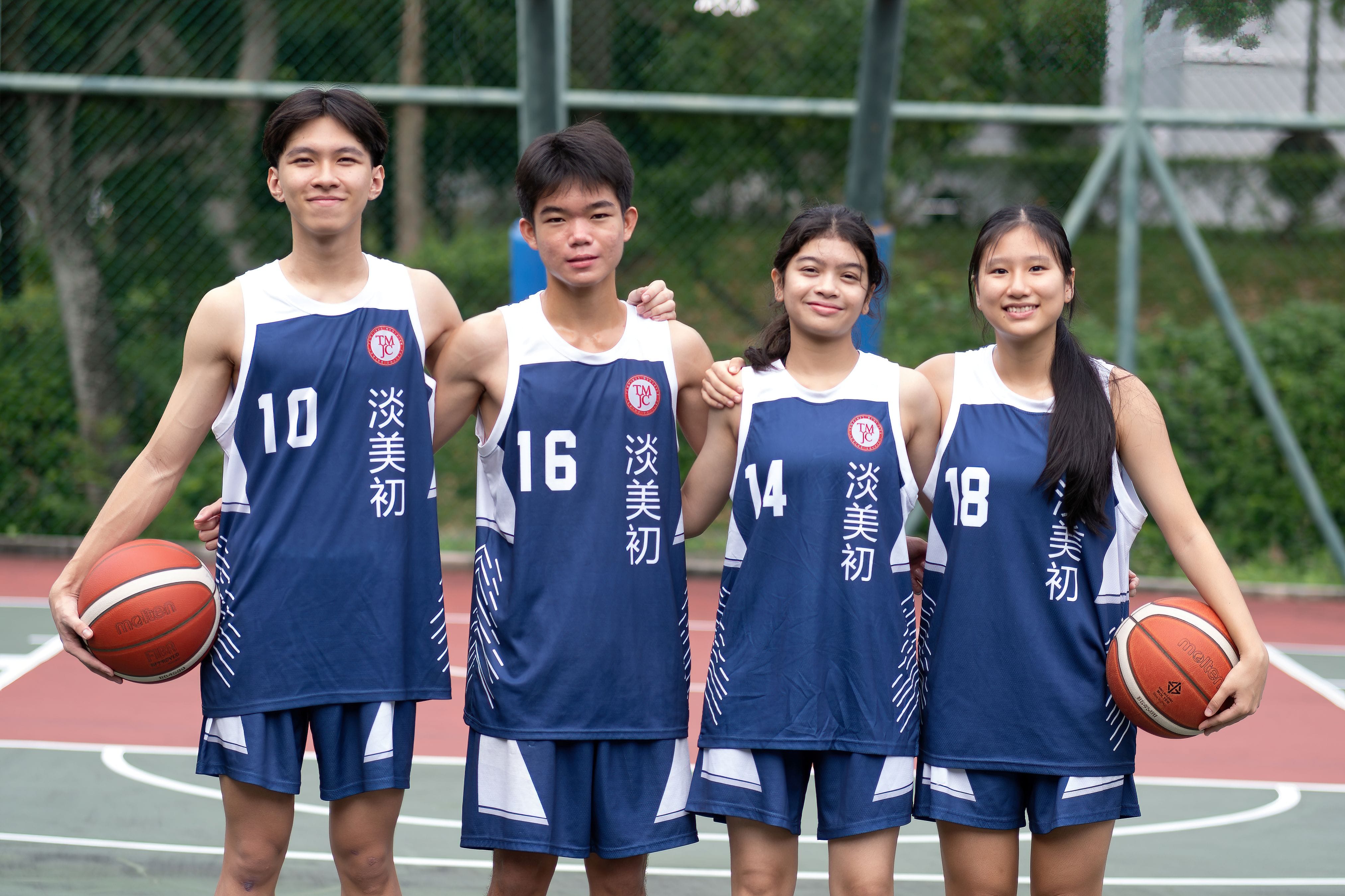 Four TMJC students in navy blue basketball jerseys (numbered 10, 16, 14, and 18) pose together on an outdoor basketball court, two holding basketballs.