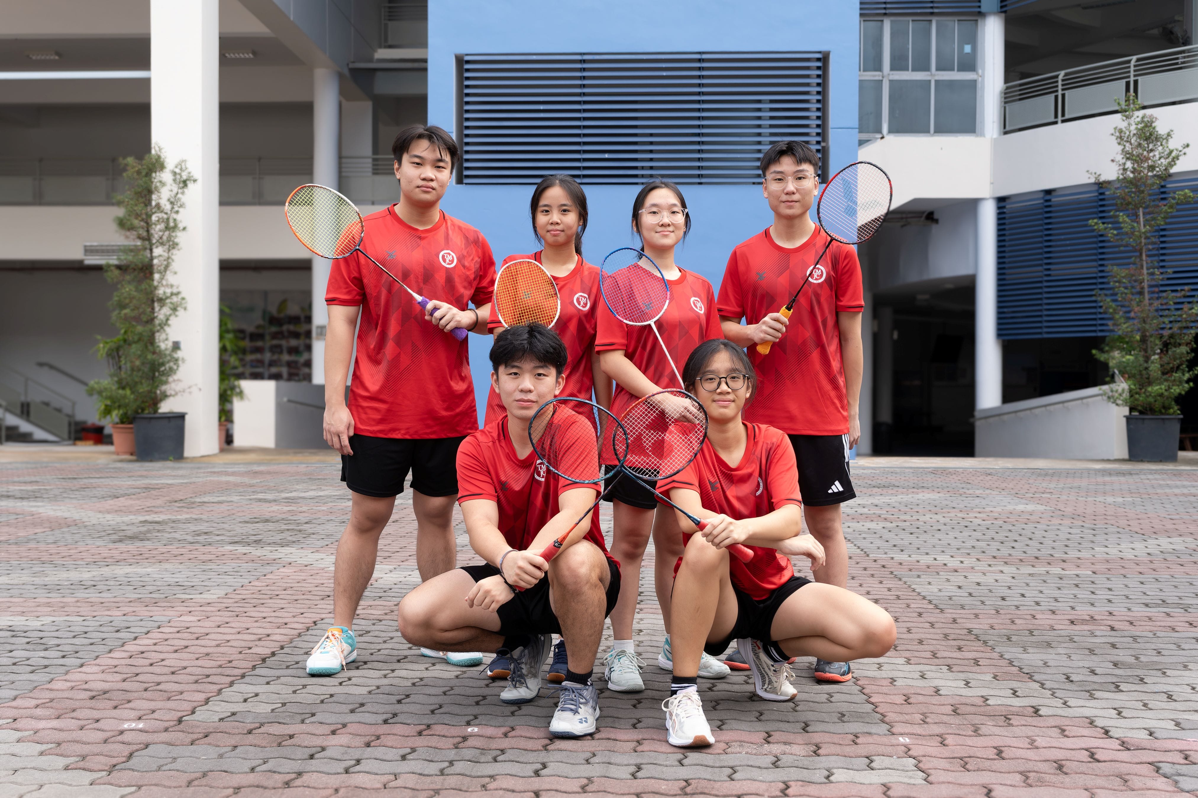 Six TMJC students in matching red jerseys pose confidently outdoors, each holding a badminton racket, with four standing and two kneeling in front.