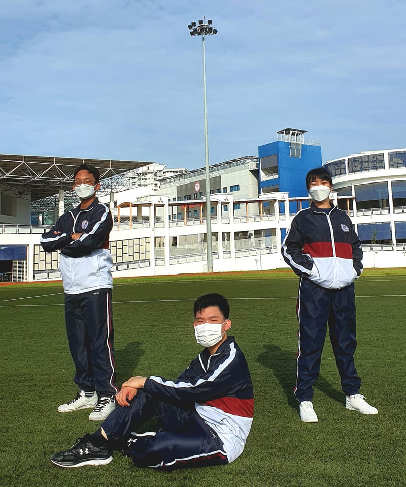 Three students in matching navy tracksuits and face masks pose on a green sports field with stadium buildings behind them.