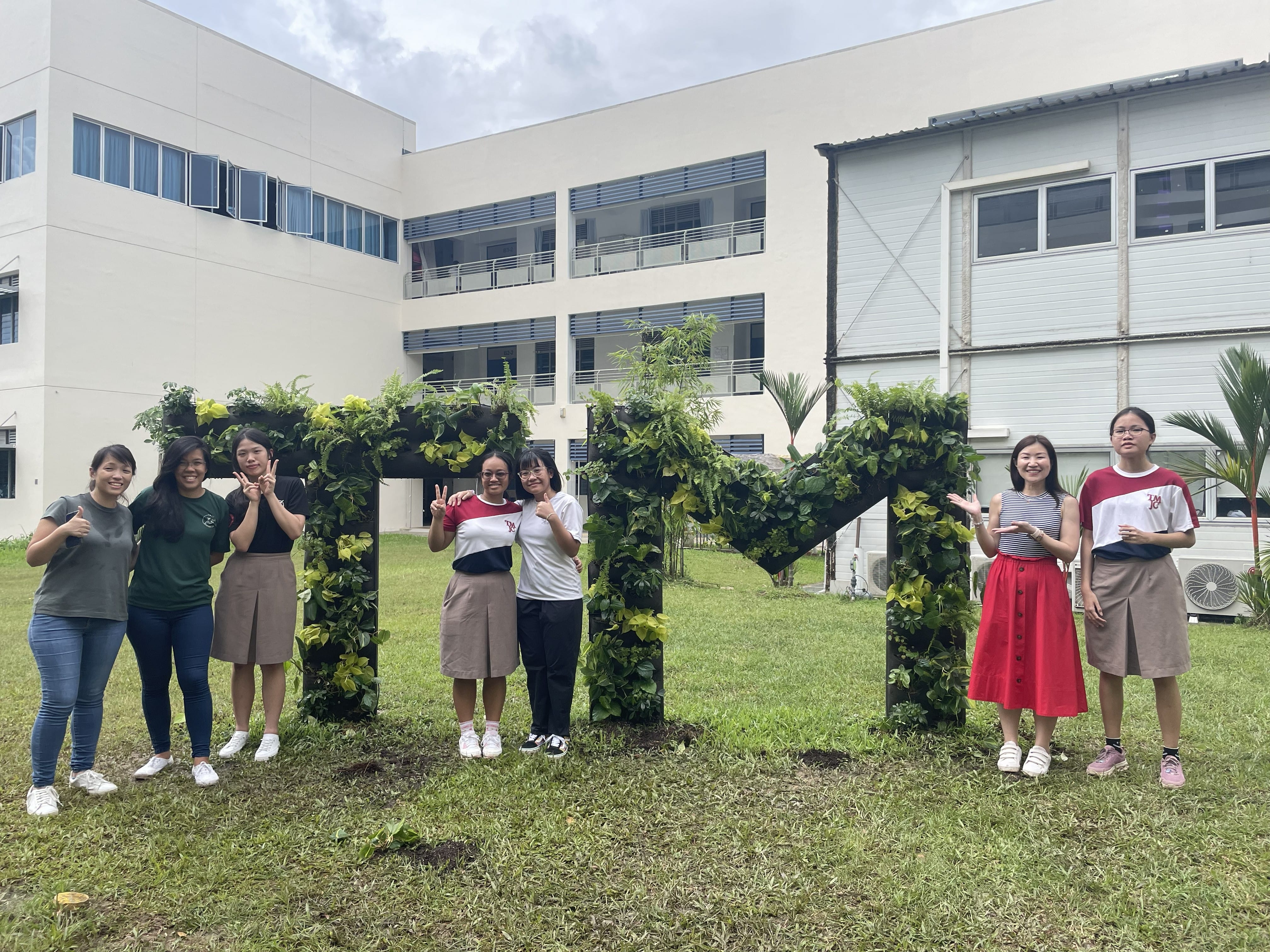 Seven students pose happily in front of large letter sculptures spelling "TM," covered in lush green climbing plants, on the school grounds. School buildings are visible in the background.