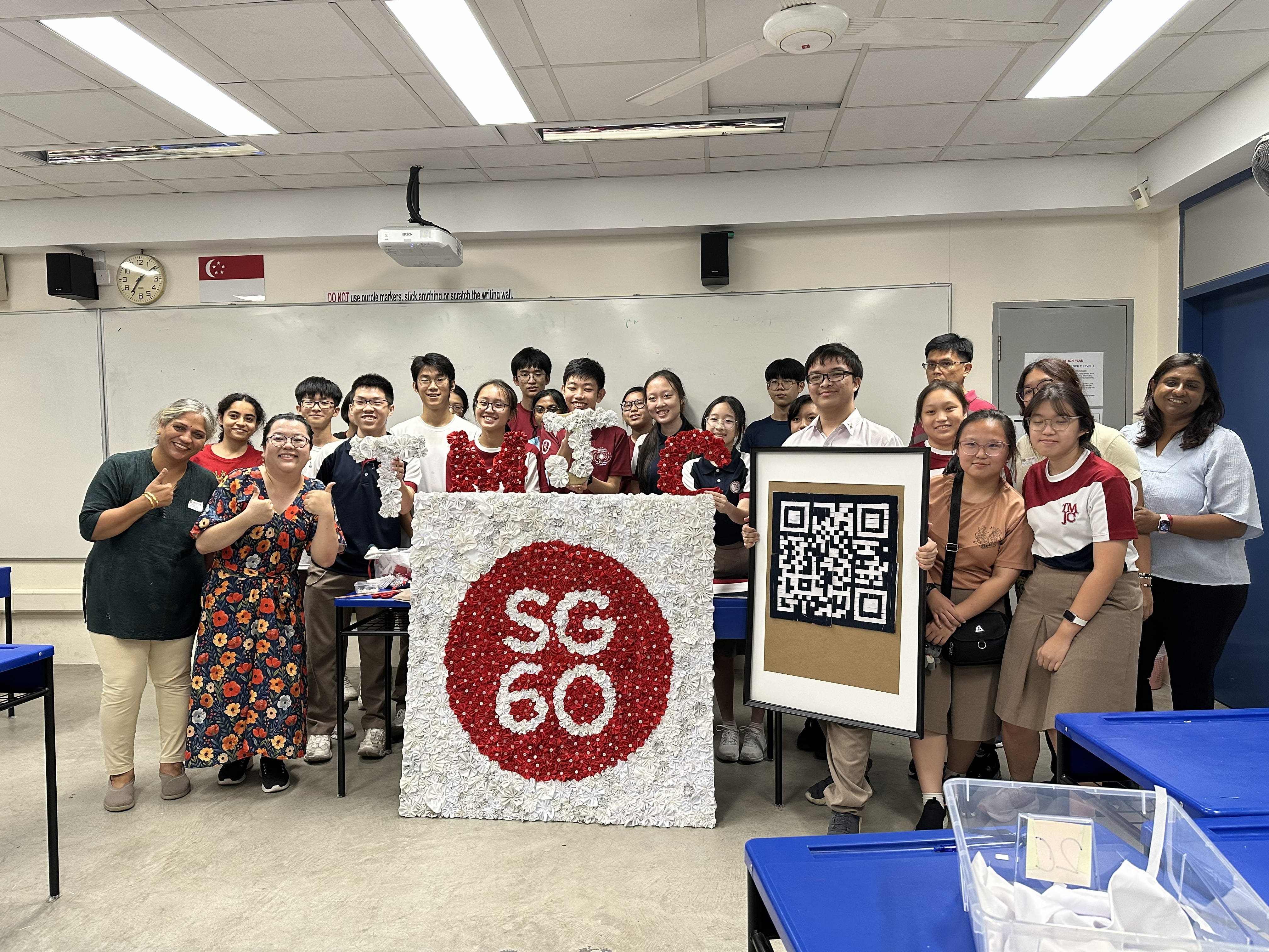 A large group of students and teachers pose cheerfully in a classroom, holding up large letters spelling "TM" and a handcrafted artwork featuring the "SG60" logo made from red and white paper rosettes. One student holds a framed QR code. A Singapore flag is visible on the wall behind them.