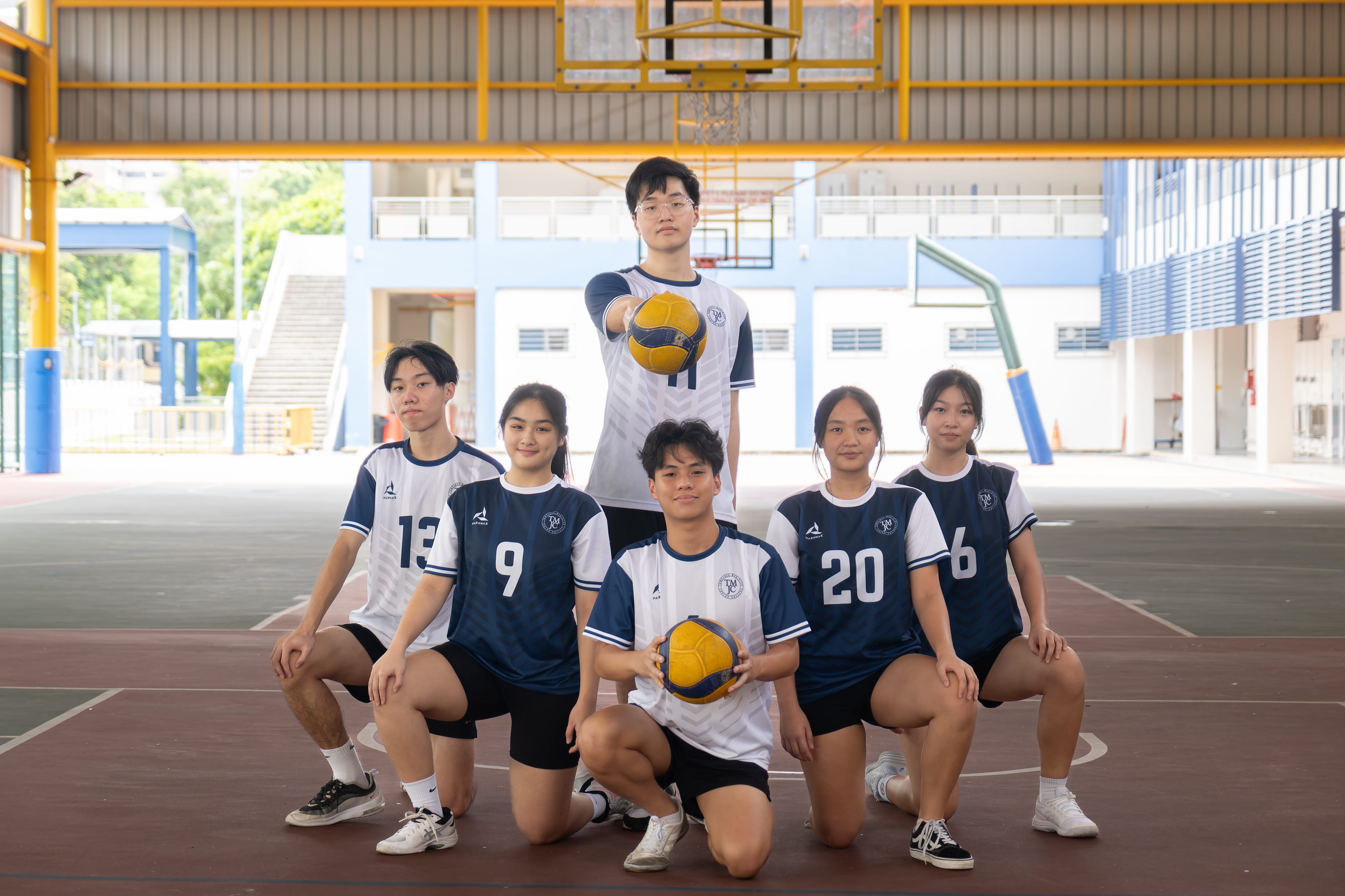Six TMJC volleyball players in navy blue and white jerseys pose together on an outdoor court, with two holding volleyballs.