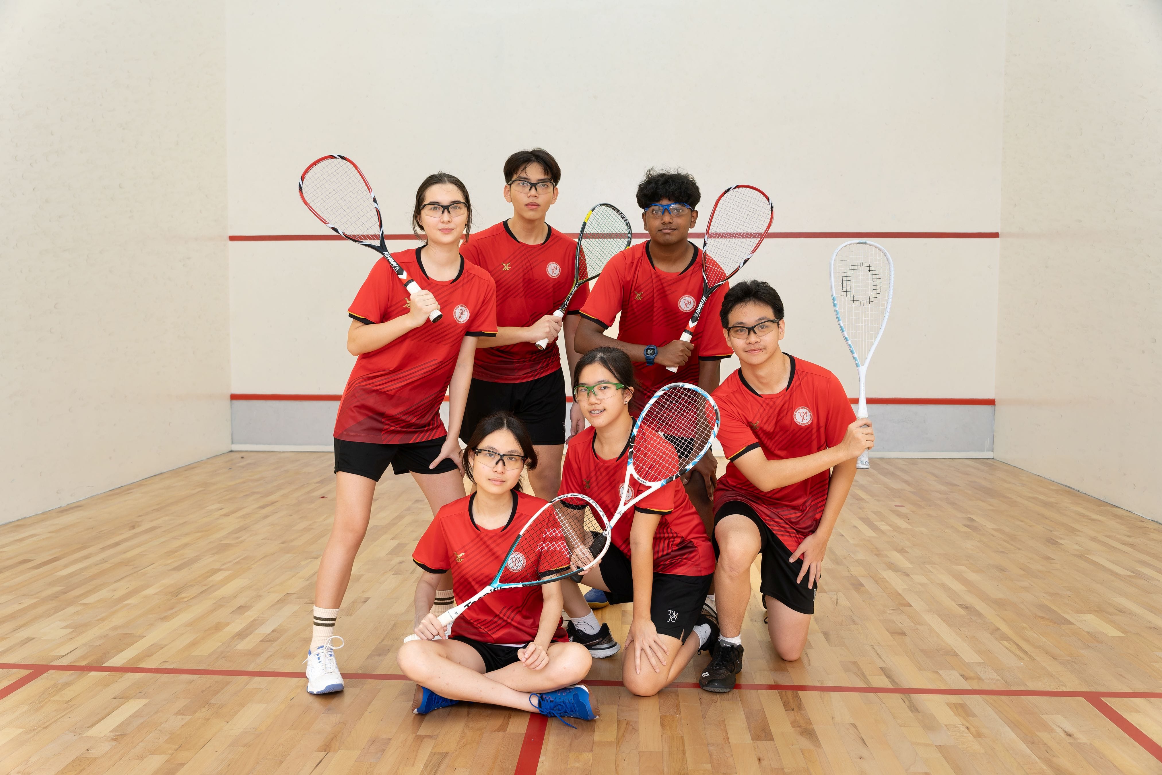 Six students in red TMJC sports jerseys pose together on an indoor squash court, holding squash rackets, arranged in two rows with three standing and three crouching in front.
