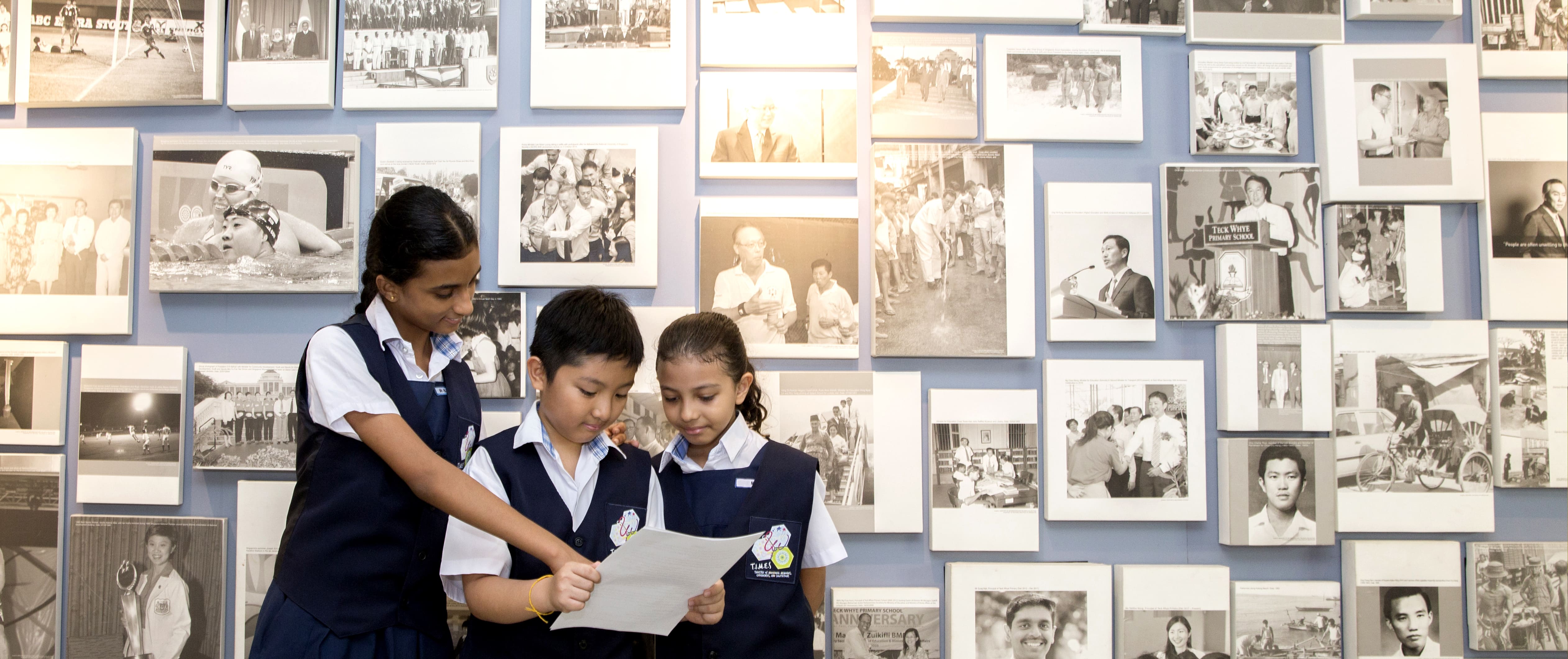 Three children in school uniforms read a paper together in front of a blue wall covered with framed black-and-white photos.