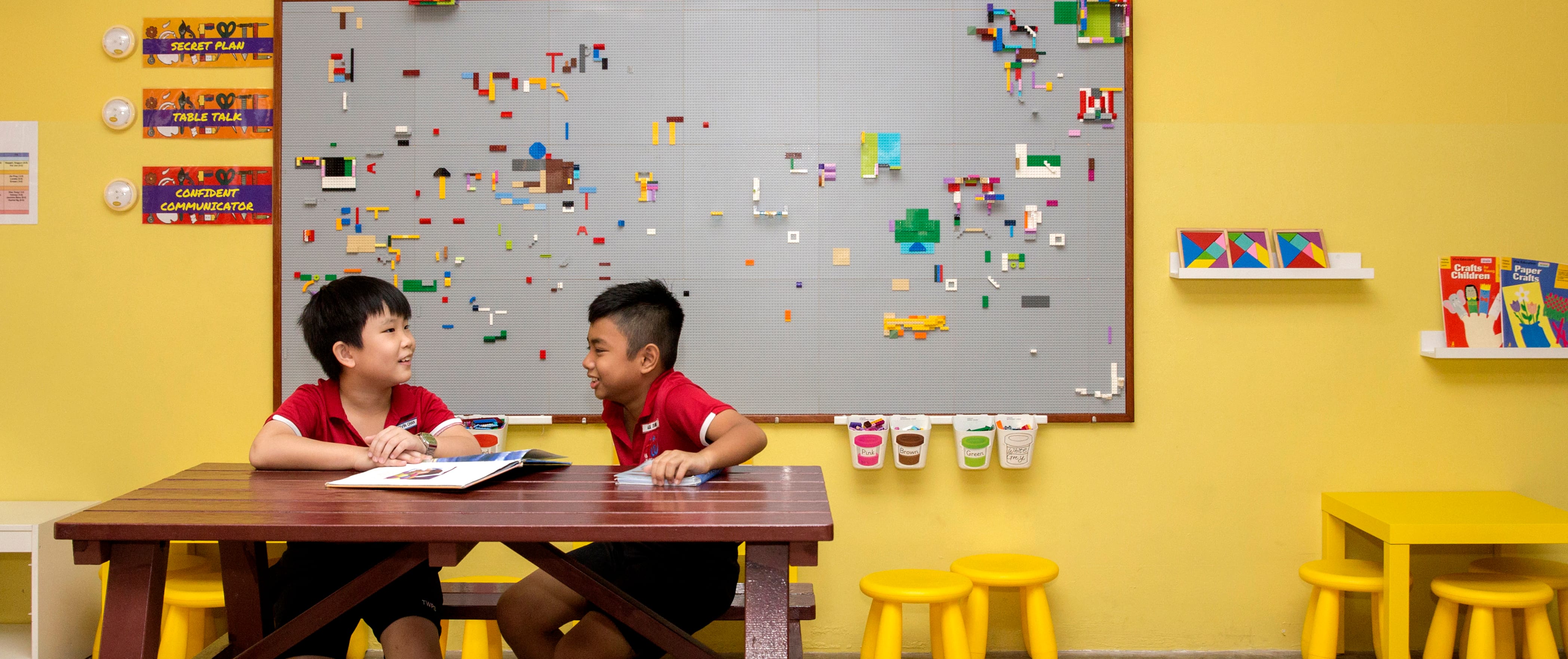 Two students seated at a table, discussing ideas over an open book in a classroom.