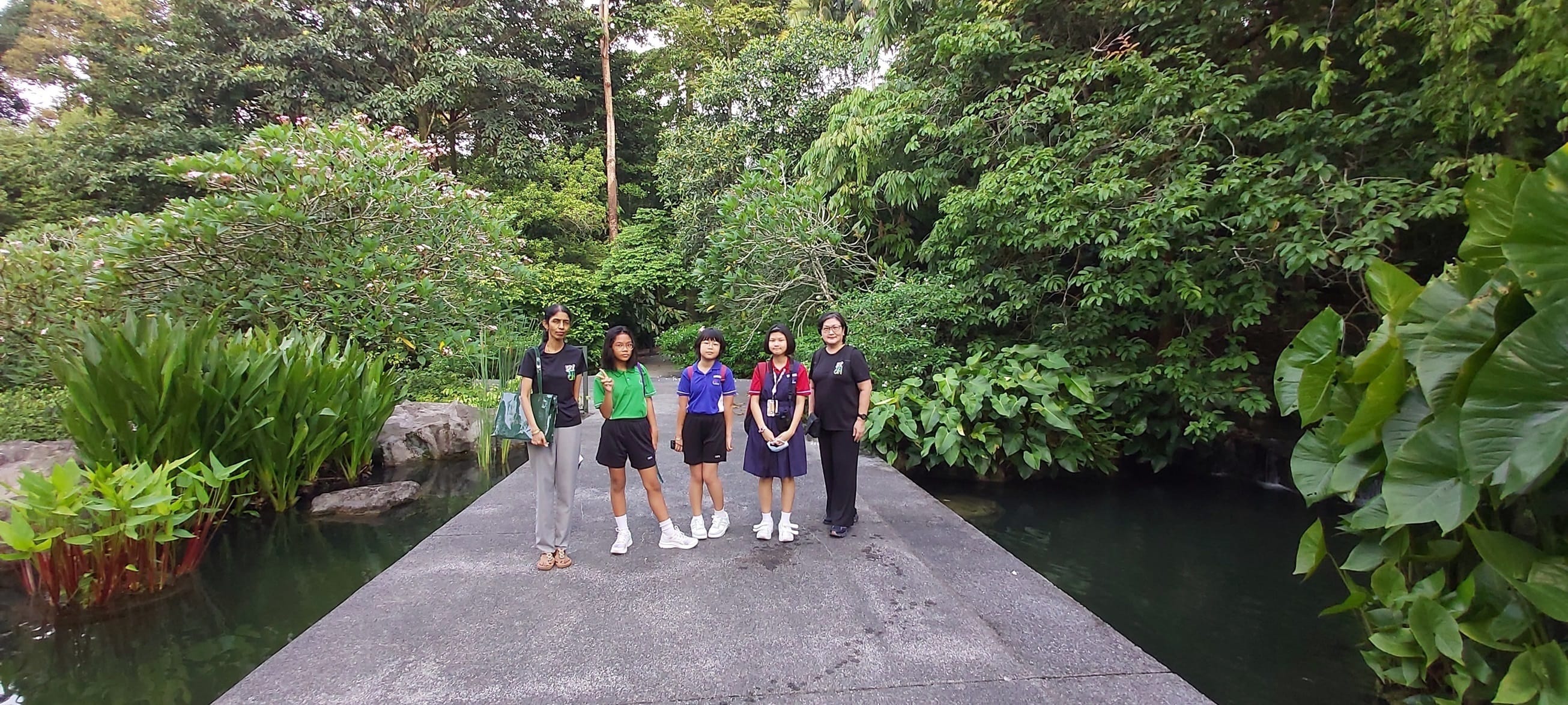 Green club students and teacher pose on a stone bridge over water, surrounded by lush green trees and tropical plants.