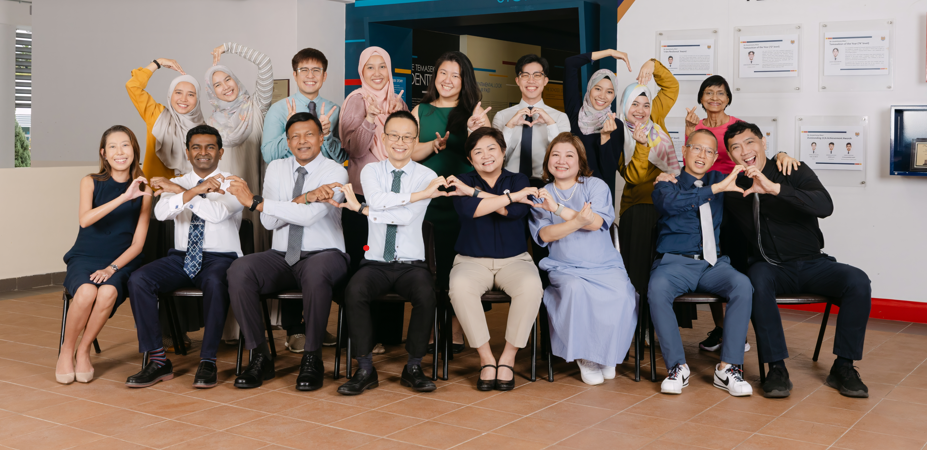 A group of about 18 people pose in two rows before a blue "Our Temasek Story" display board.