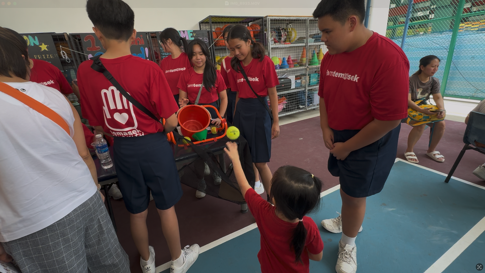 Teens in red Temasek shirts supervise a young child reaching for a ball at an indoor games booth.