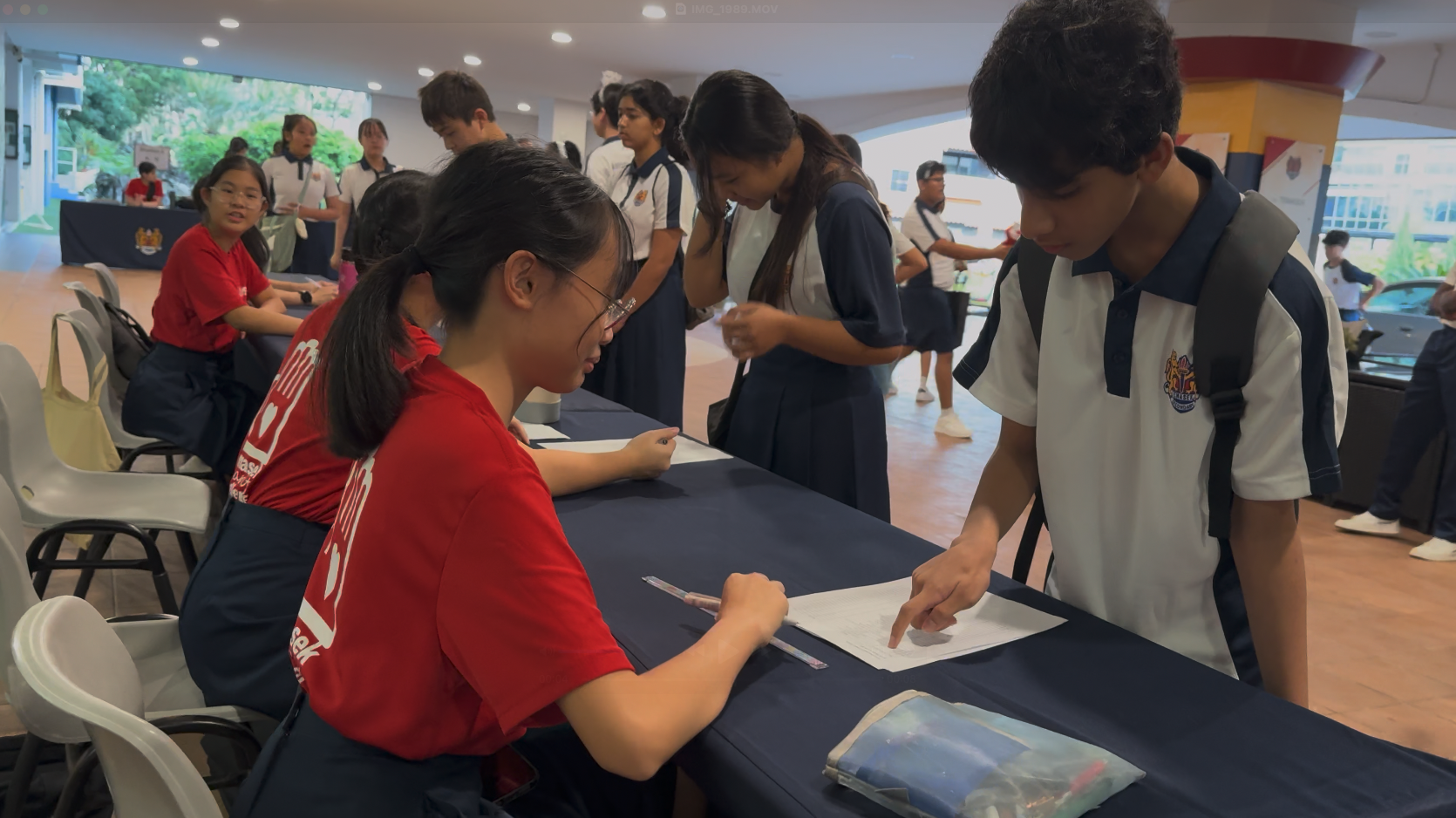 Students in school uniforms gather at tables with papers during what appears to be a school registration or event.