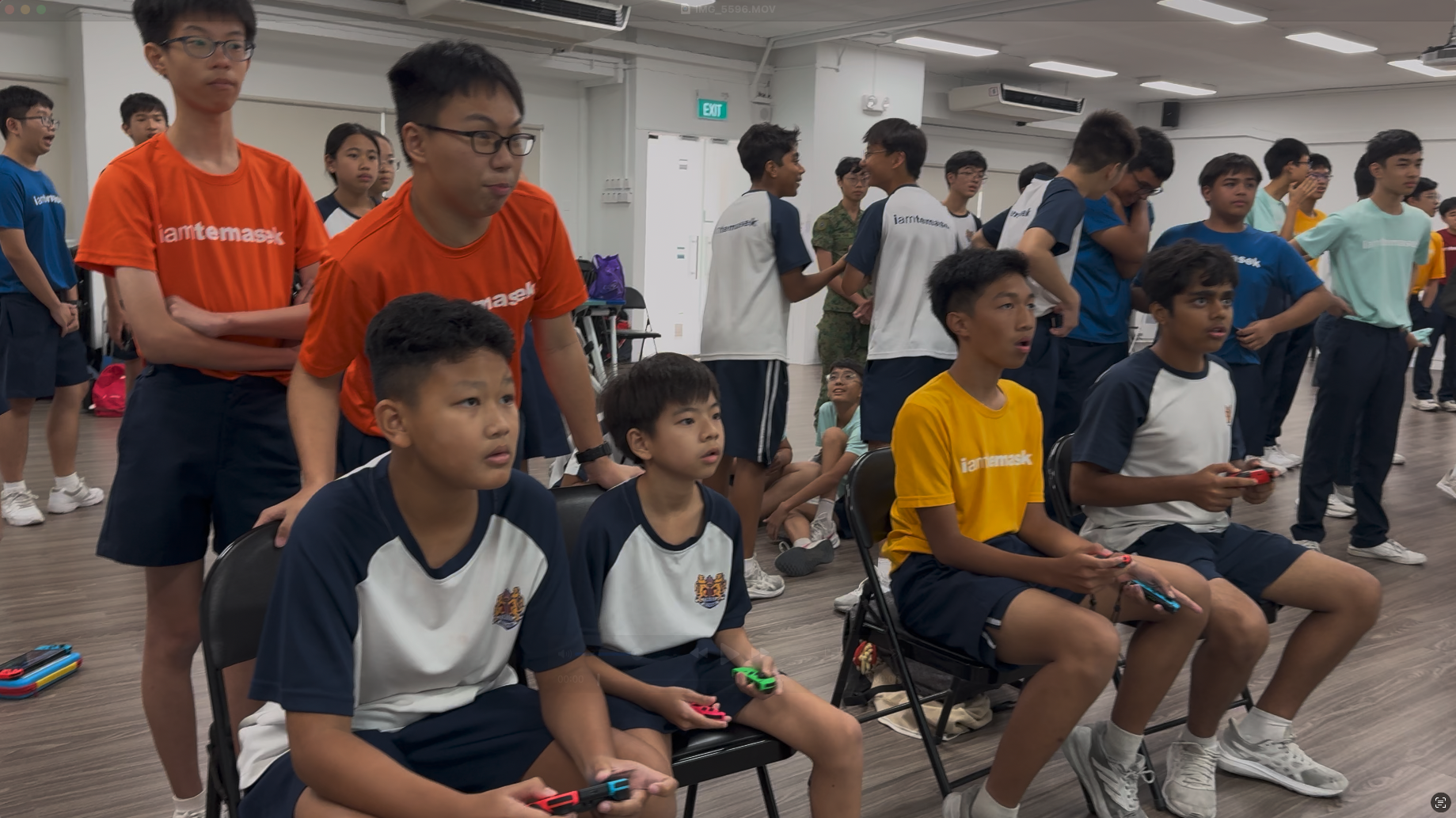 Students in school uniforms sit and stand in a room, holding Nintendo Switch controllers while others watch.