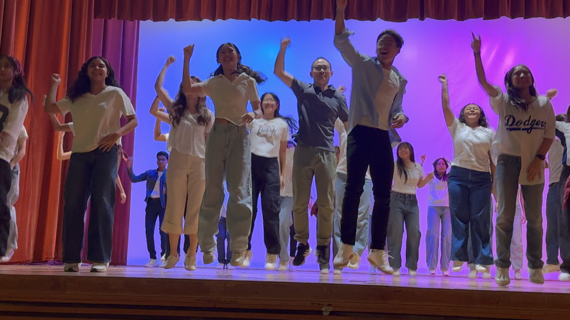 A large group of teens dance and jump on a stage with red curtains and blue-purple lighting backdrop.