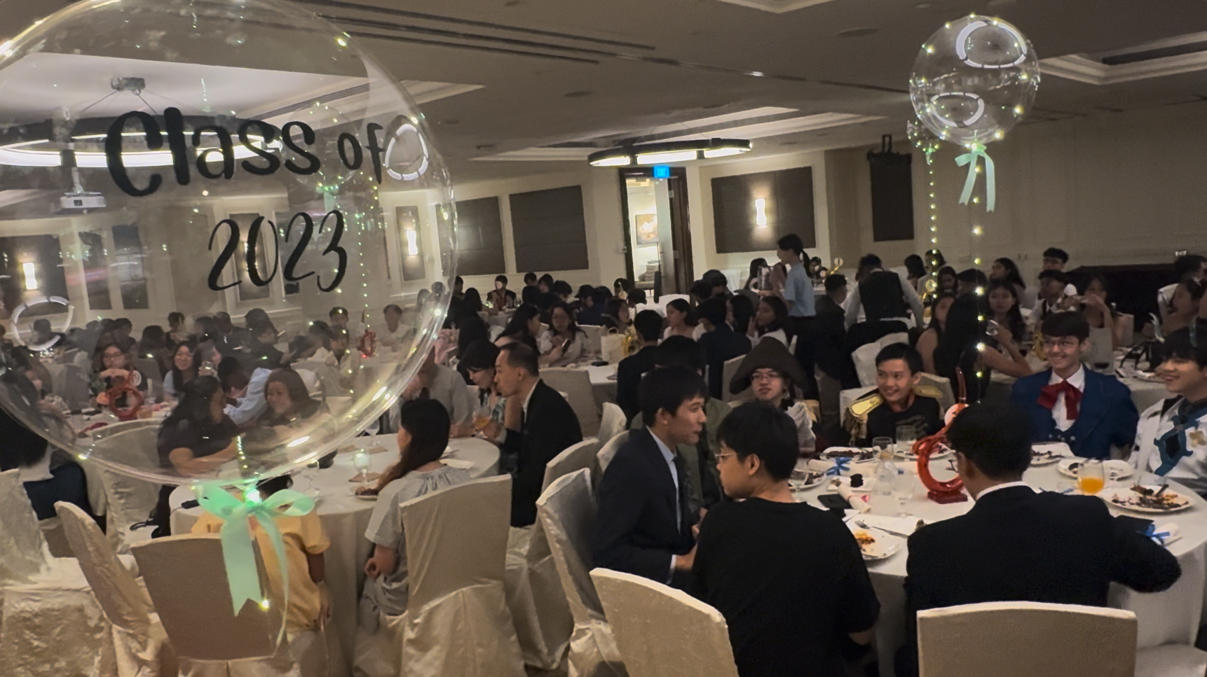 Formal banquet hall filled with students at round tables, with a large clear balloon reading Class of 2023 in the foreground.