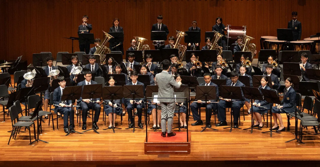 A conductor in a grey suit leads a large youth wind band in dark uniforms on a wooden concert stage.
