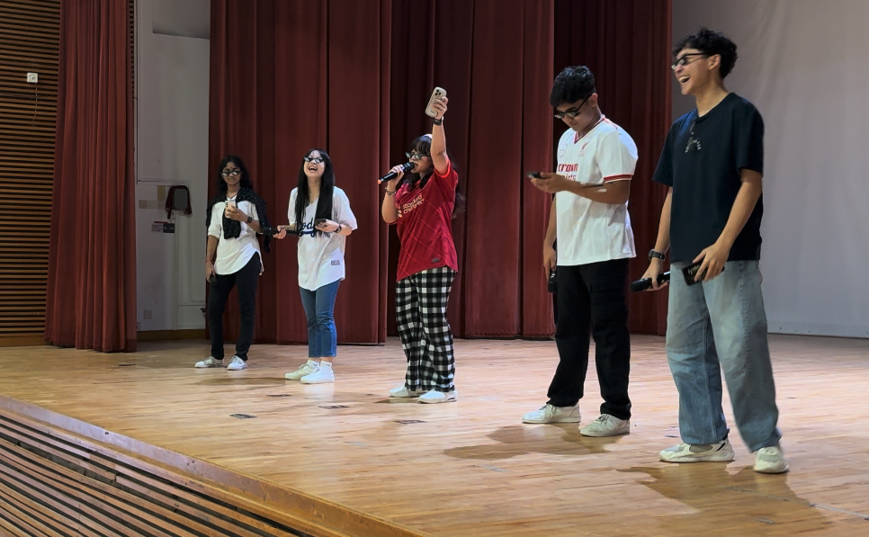 Five teenagers stand on a wooden stage with red curtains, one holding a microphone while others hold phones.