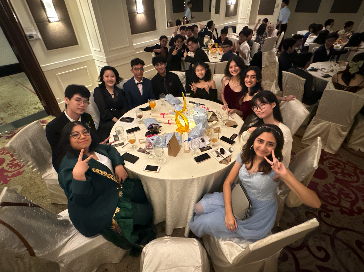 A group of dressed-up teens smile and pose with peace signs at a round banquet table in a formal hall.