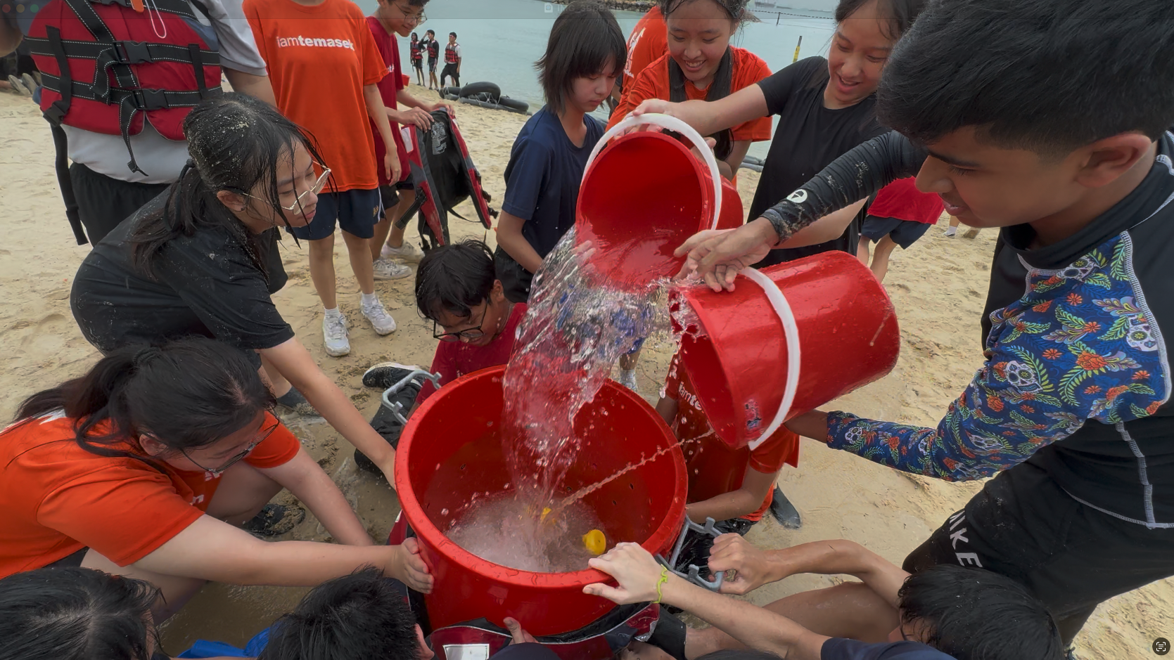 A group of young people on a beach pouring water from red buckets during an outdoor team activity.