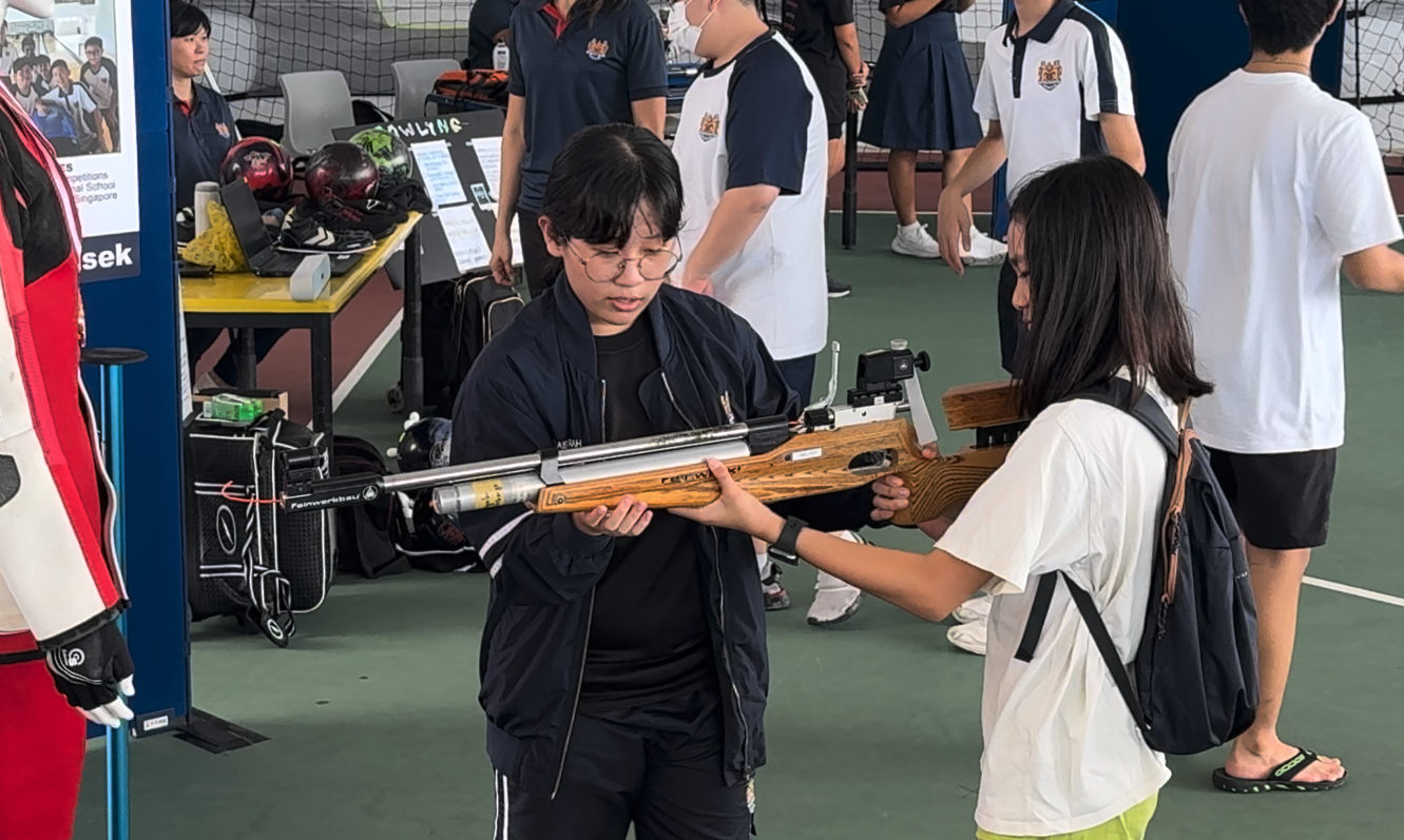 Two students handle a wooden air rifle at a school sports exhibition with other students and display tables in the background.