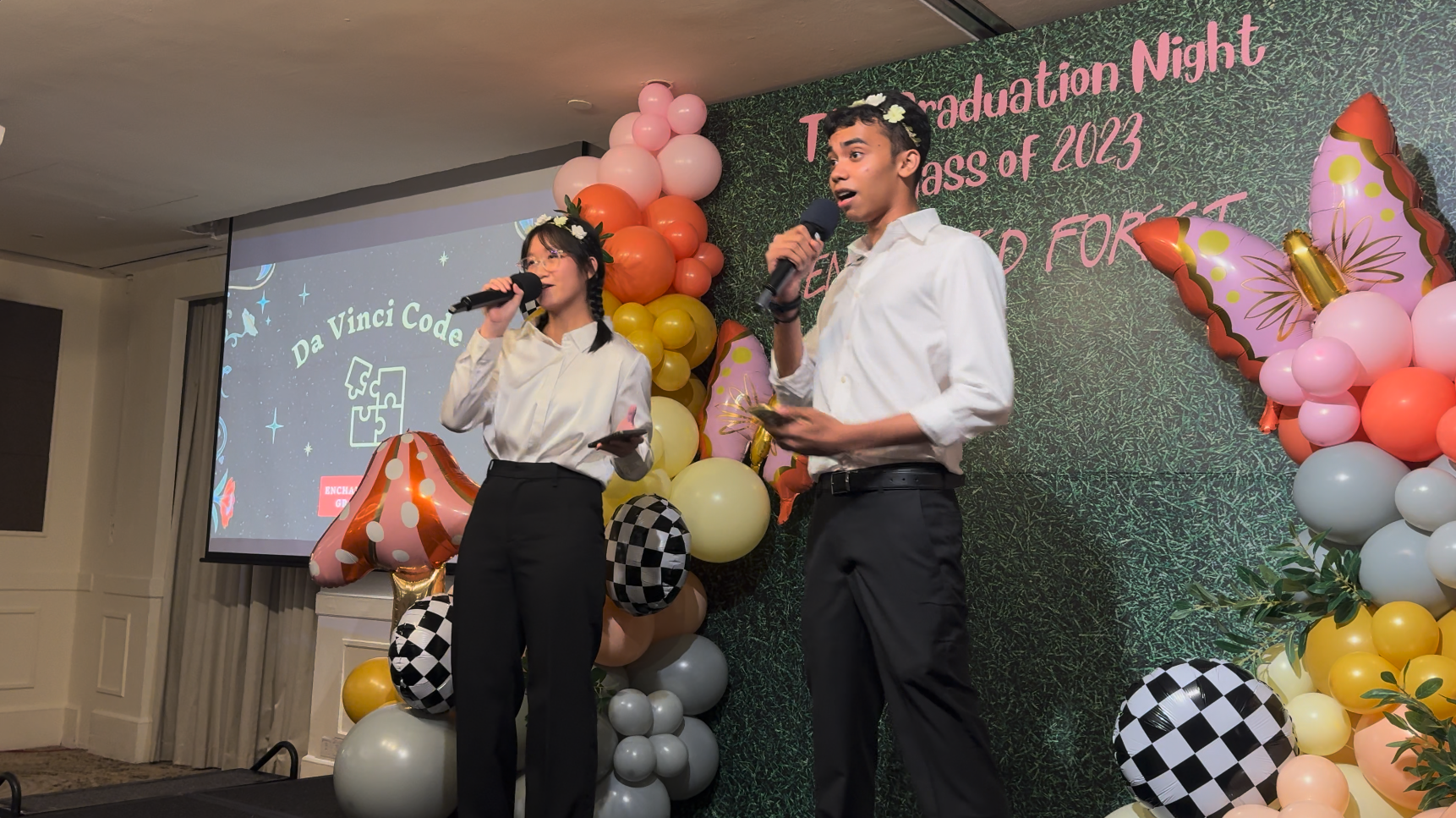 Two students holding microphones on stage at a Class of 2023 Graduation Night event with colorful balloon decorations.