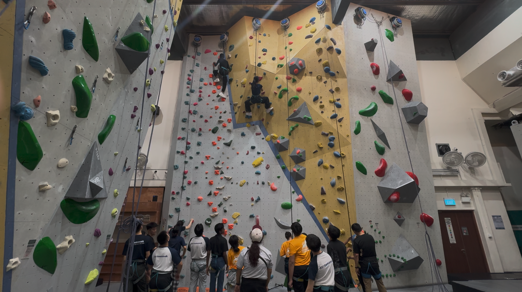 A group of people stand at the base of a tall indoor climbing wall while two climbers ascend using colorful holds.