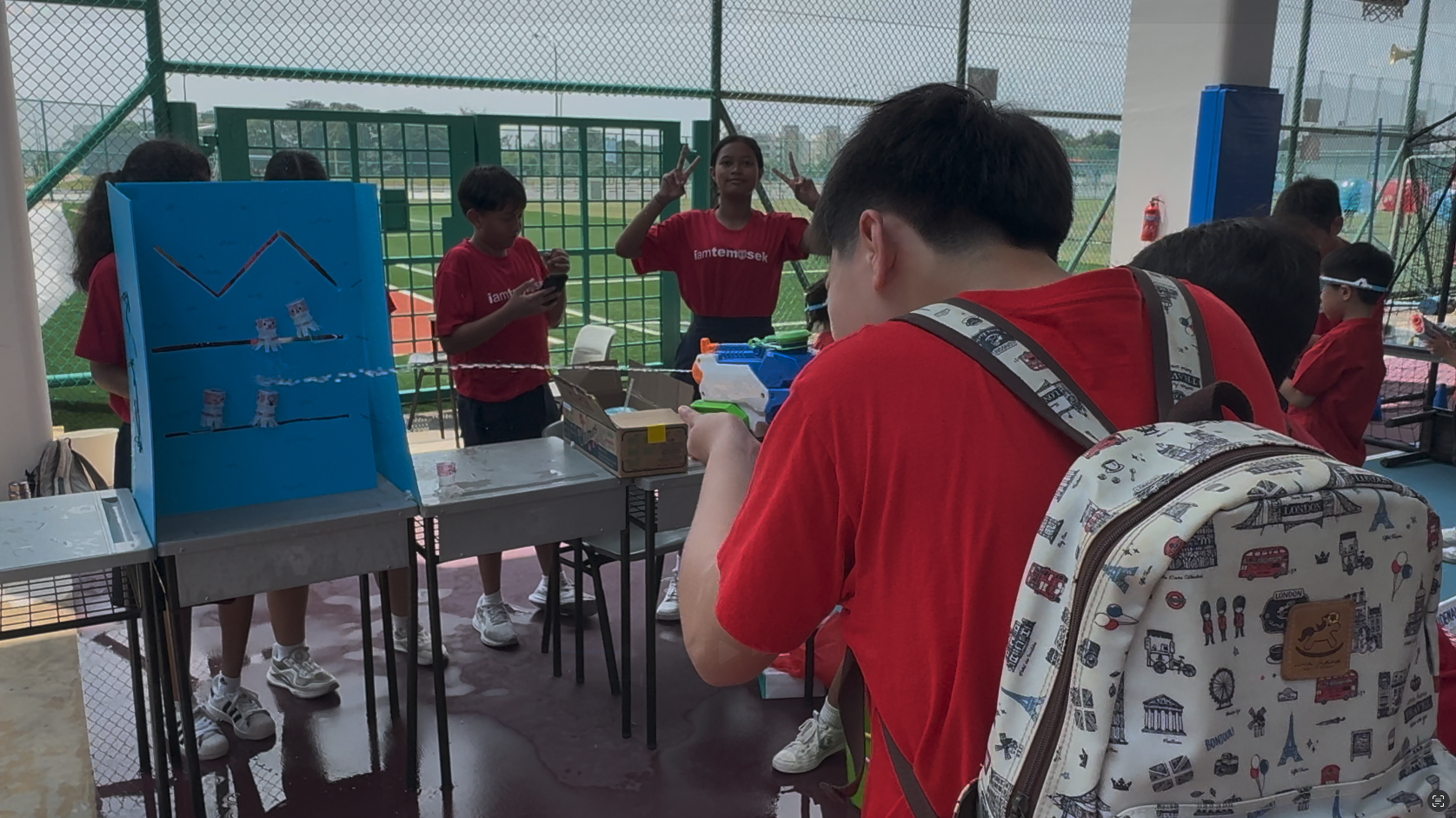 Students in red shirts play a carnival shooting game with a toy gun aimed at a blue cardboard target display.