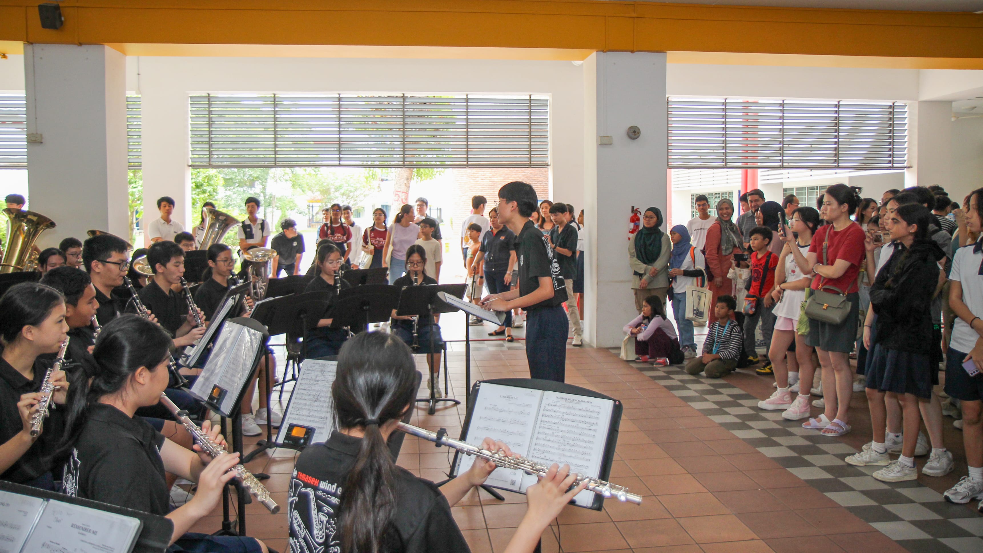 Student musicians play flutes and brass as a conductor leads, while a crowd watches in a covered walkway.