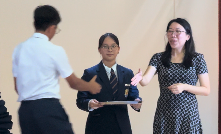 A woman in a patterned dress and a student in a blazer present a certificate to a boy in a white shirt.