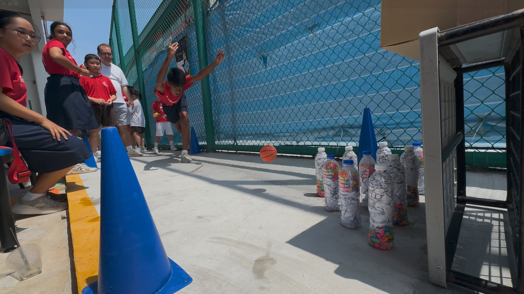 Child rolling a basketball toward plastic bottle pins in an outdoor bowling game while others watch.