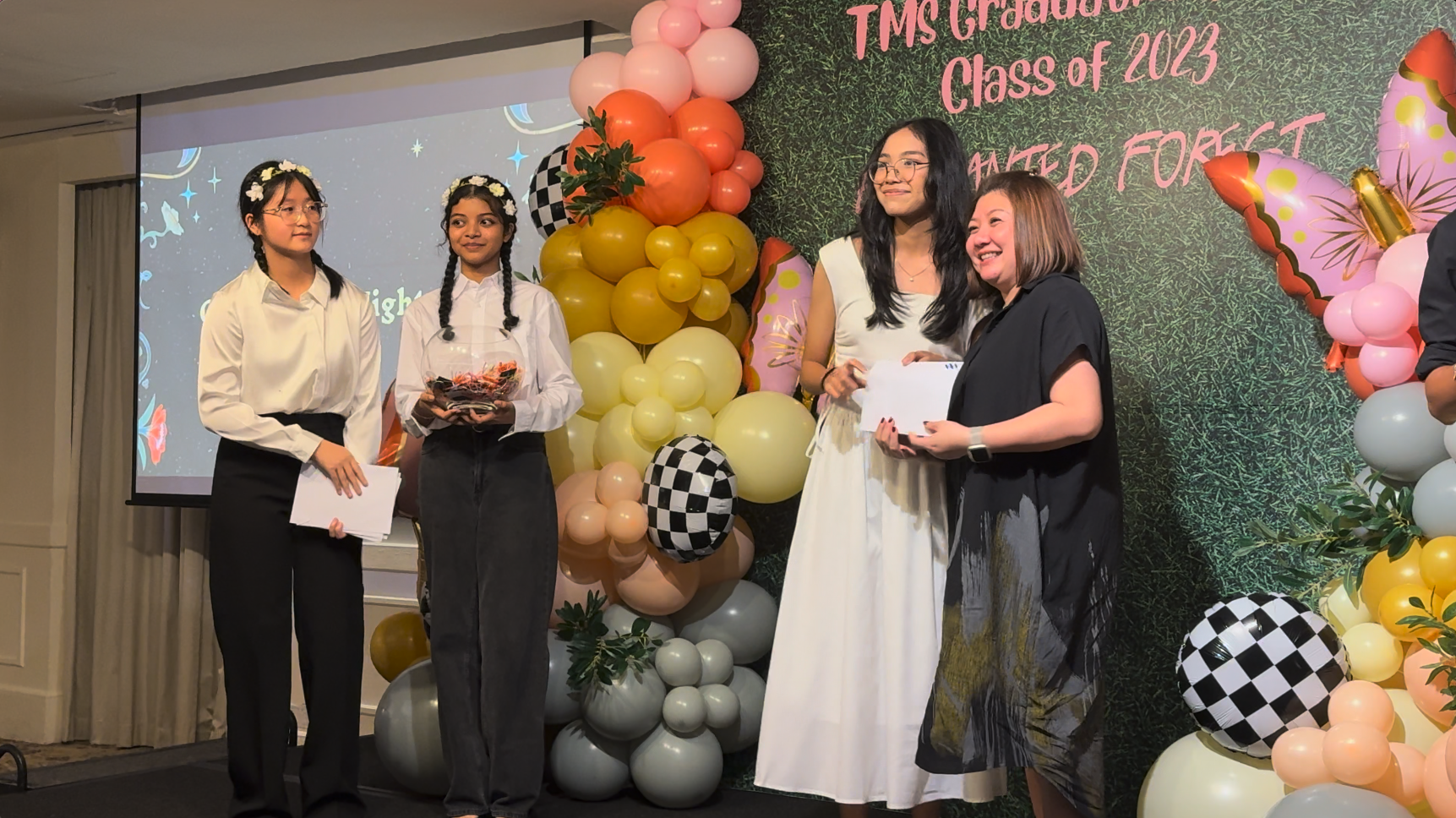 Four people stand on a stage at TMS Graduation Class of 2023, surrounded by colorful balloon decorations.