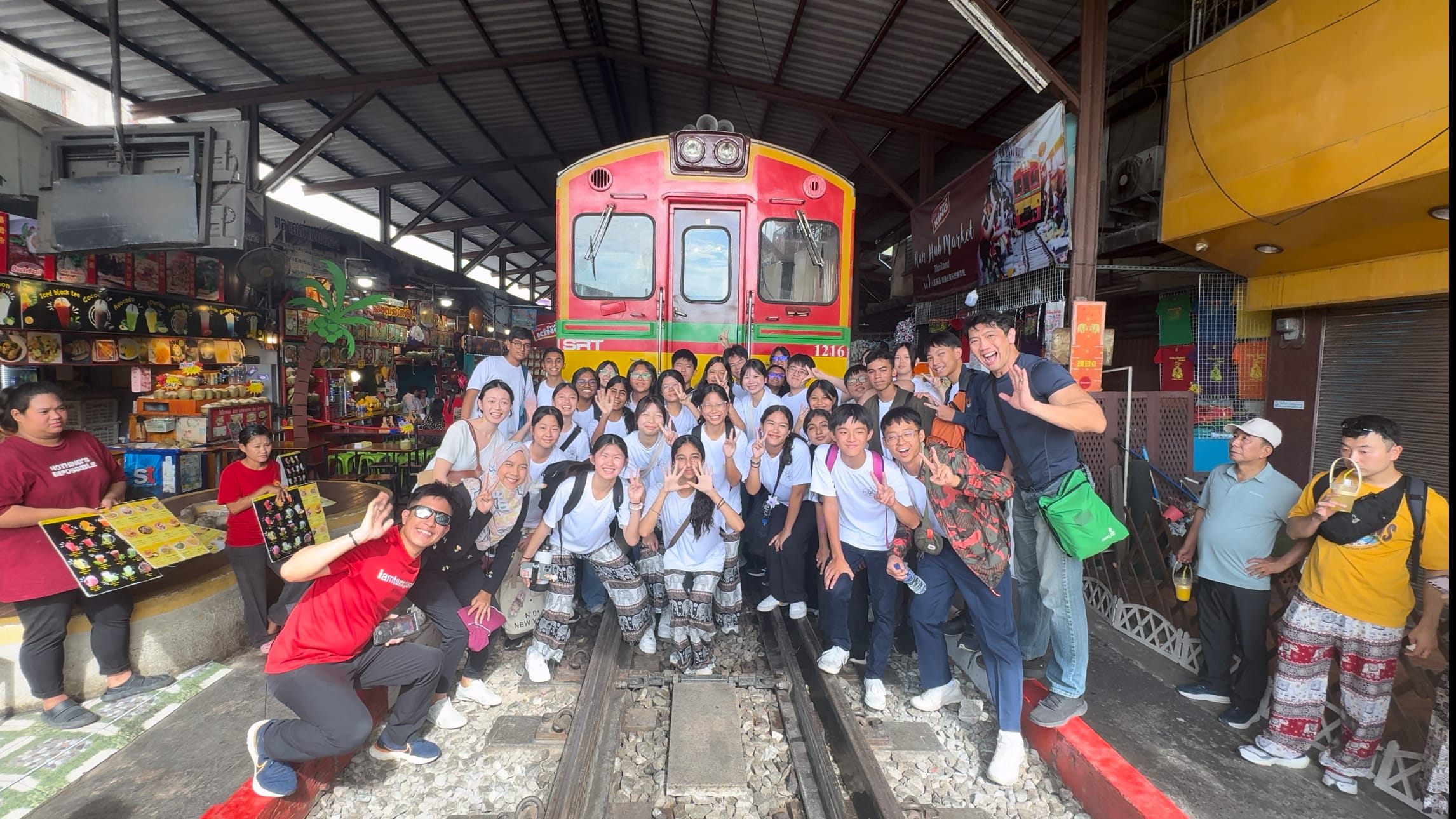 A large group of students in white shirts poses on train tracks in front of a red and yellow train at a market.