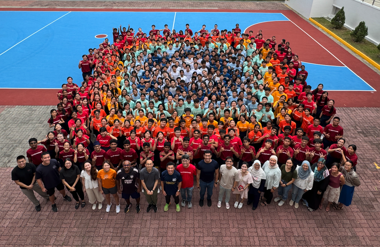 Large group of students in colorful t-shirts posing on a blue and red outdoor sports court, viewed from above.