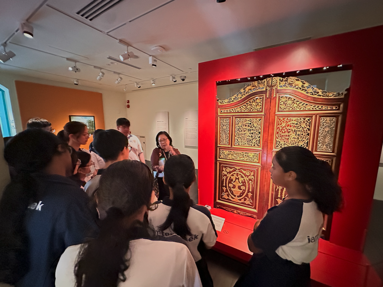 A guide speaks to a group of students in a museum gallery beside a large ornate red and gold carved wooden door.
