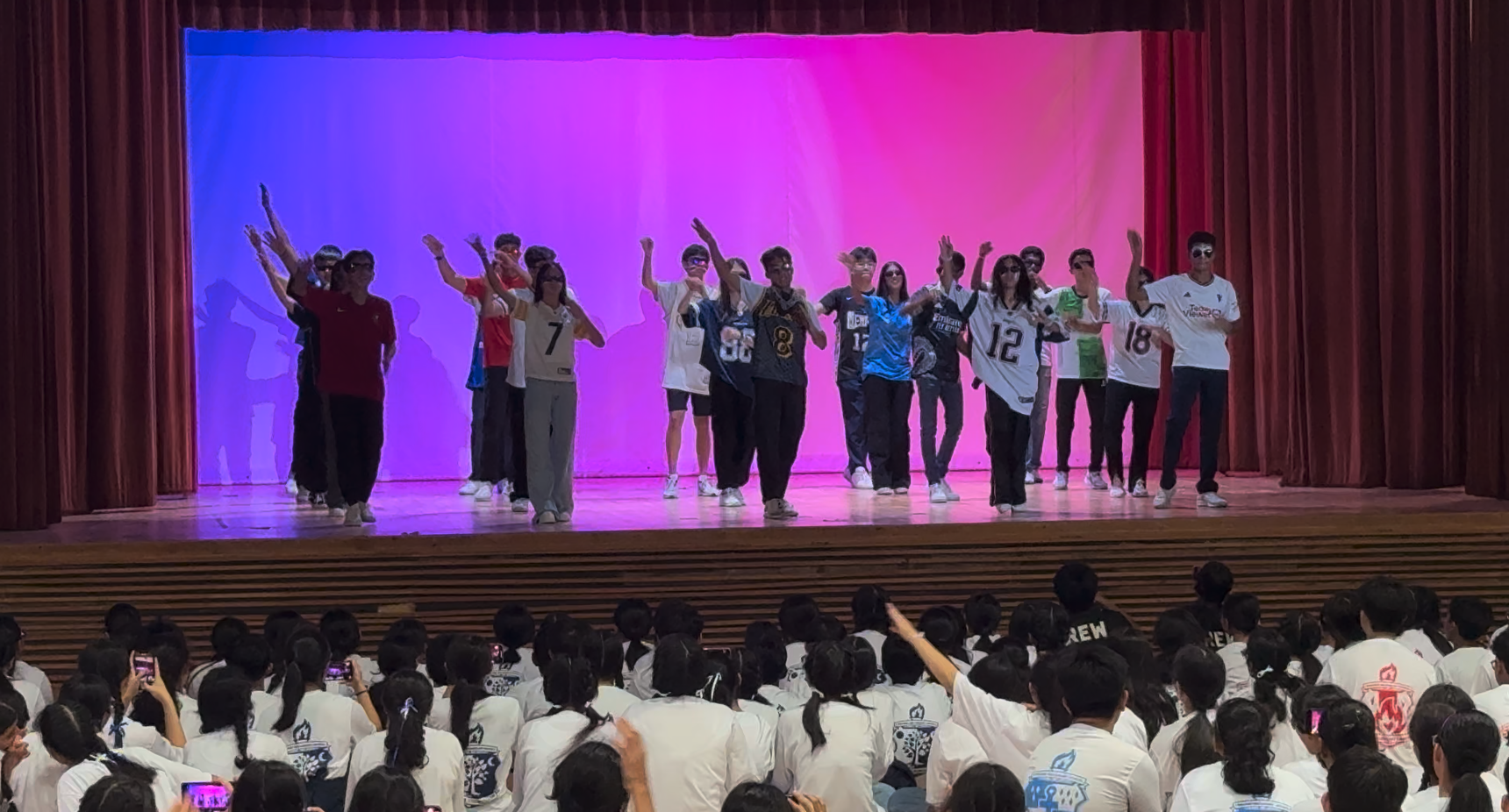 A group of teens in sports jerseys perform on a lit stage before a large seated audience in white uniforms.