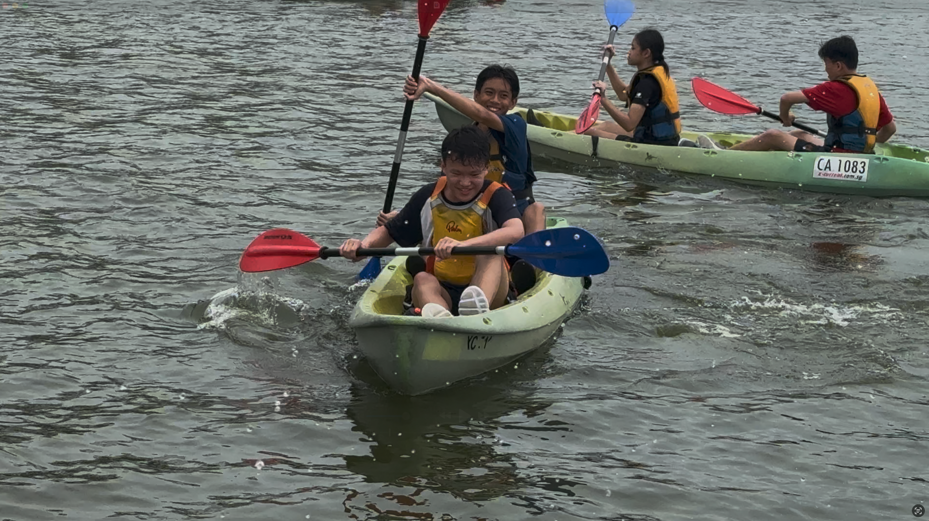 Four children wearing life jackets paddle kayaks on calm water, smiling and holding colorful paddles.
