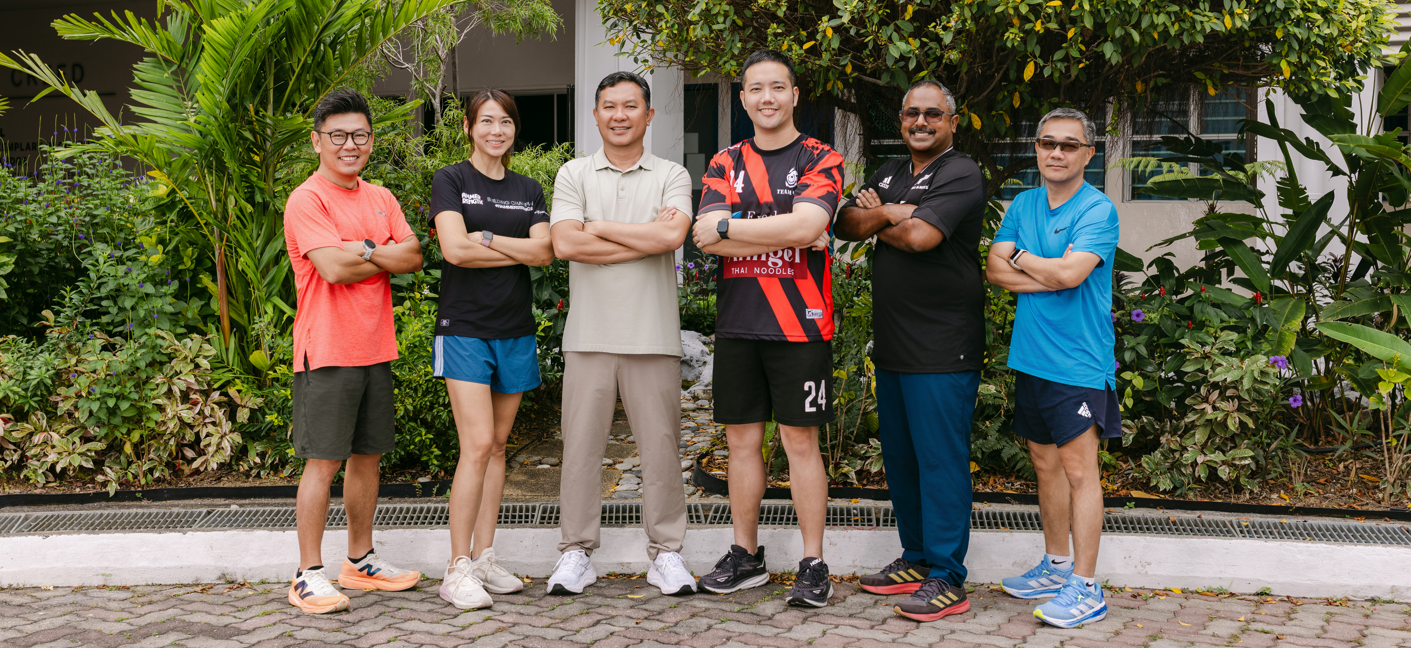 Five people in casual sportswear posing outdoors at a picnic bench surrounded by green shrubs and trees.