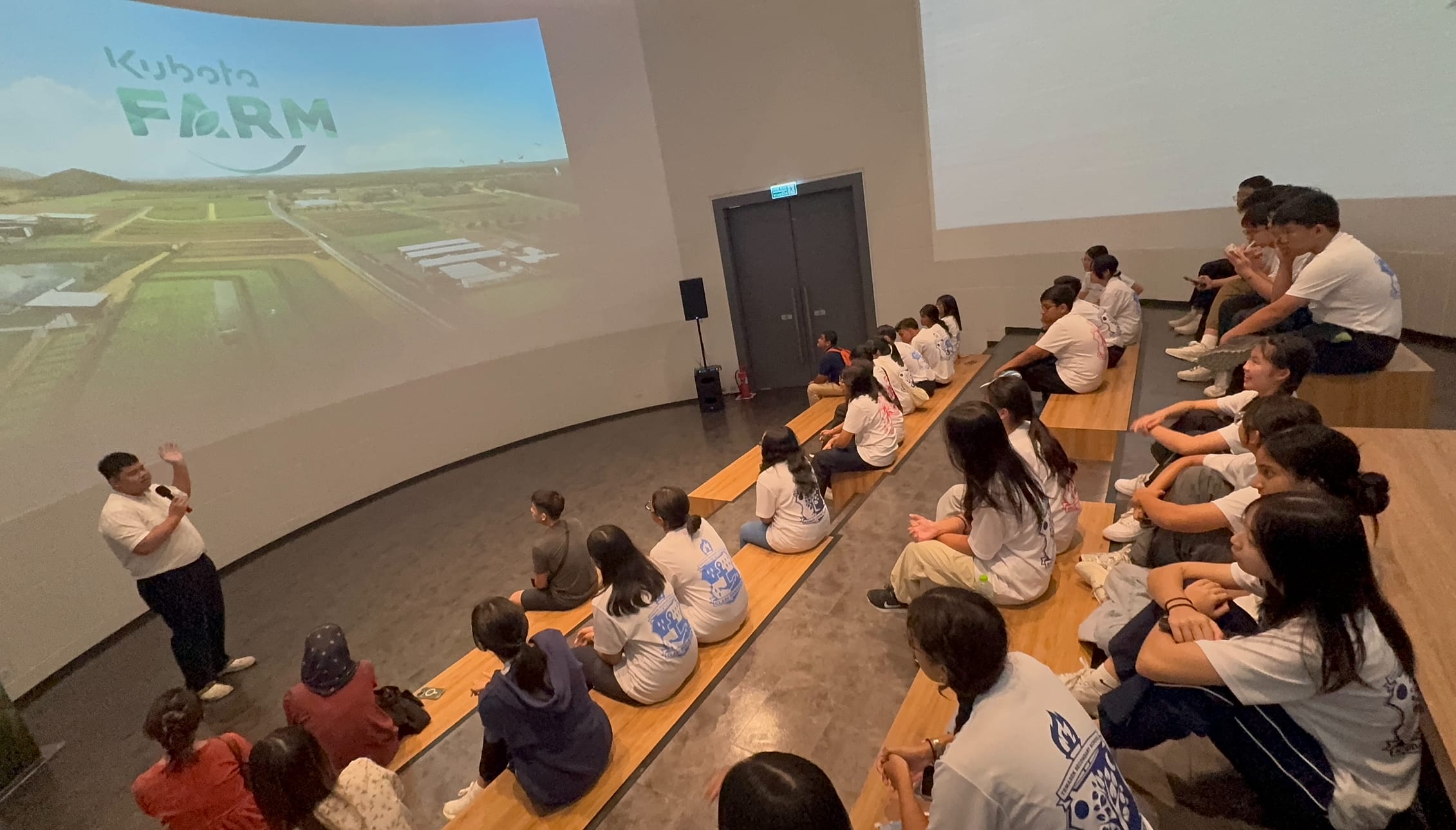 A presenter speaks to students seated on benches facing a large Kubota Farm projection screen in a modern hall.