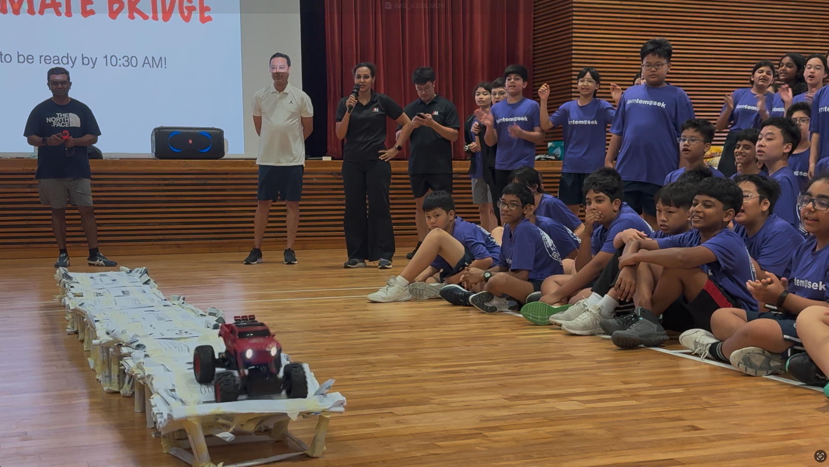 Students in blue shirts watch a red toy truck drive across a paper bridge during a school event in a hall.