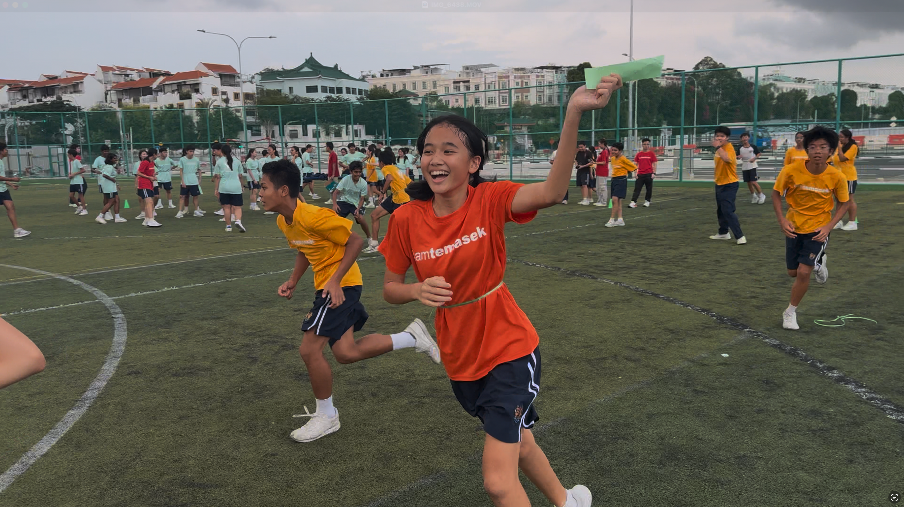 A smiling girl in an orange shirt runs on a sports field holding a green paper, with many students playing behind her.