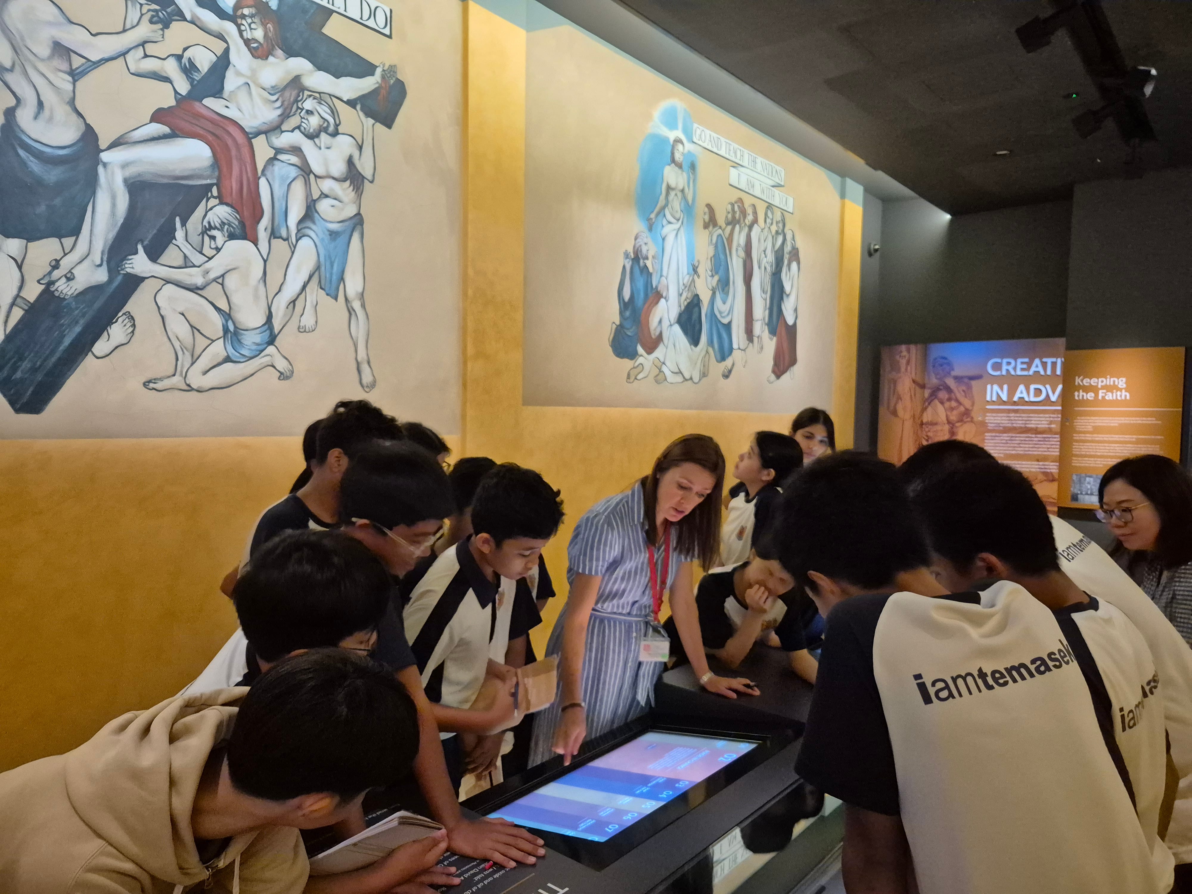 Students gather around a touchscreen display as a guide points; religious murals painted on the wall behind.